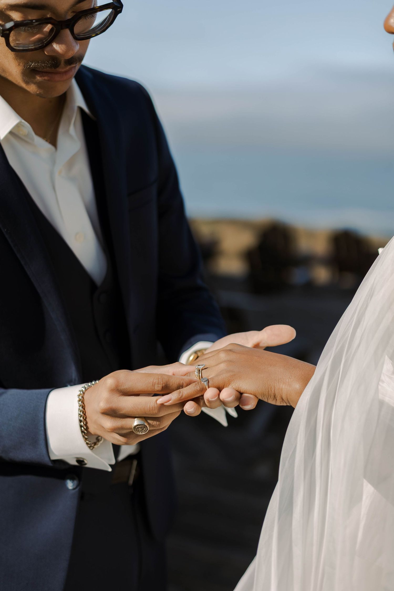 Close-up of wedding ceremony ring exchange with elegant navy suit and white dress against coastal backdrop.