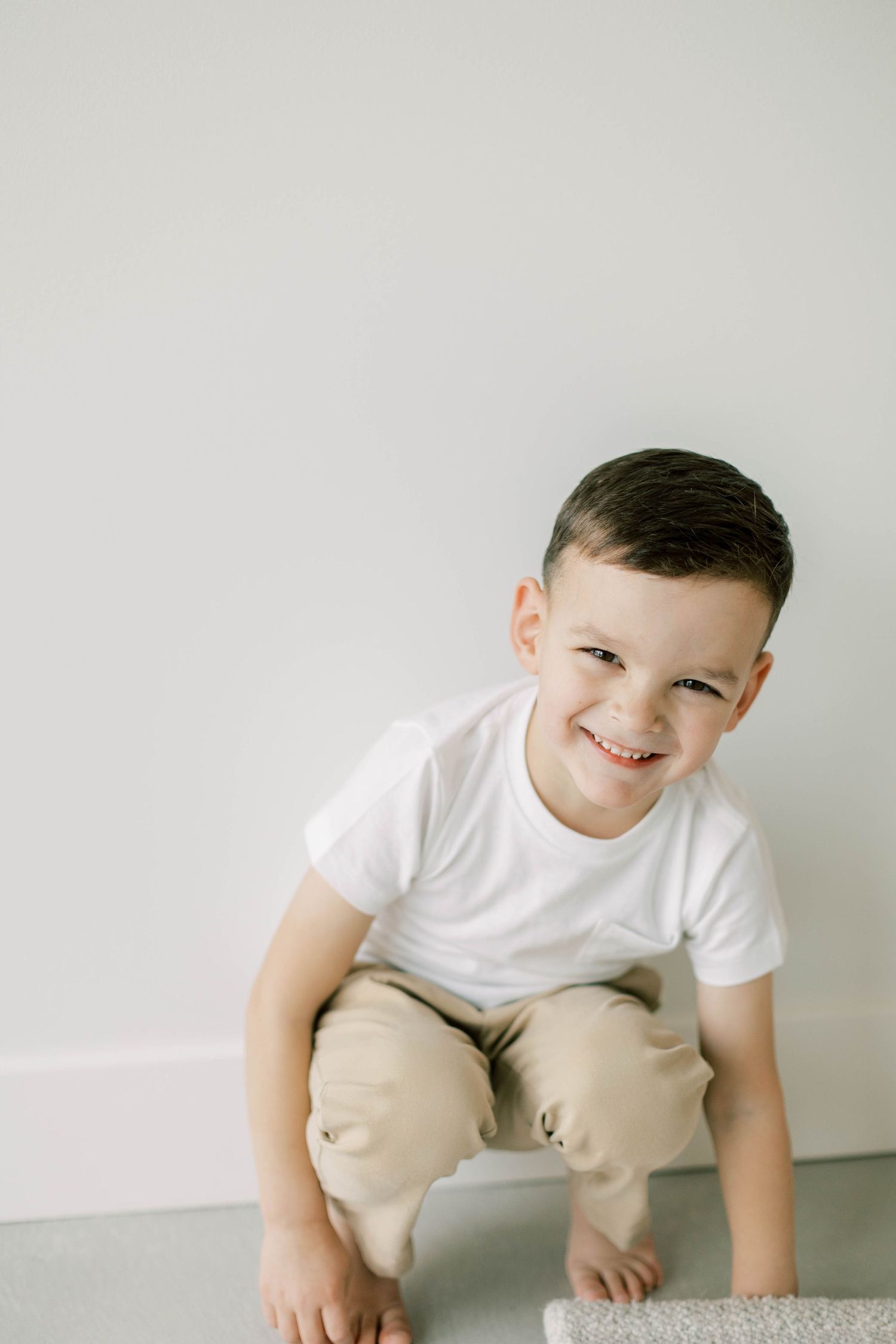 Young child in casual attire crouching and smiling against white wall background.