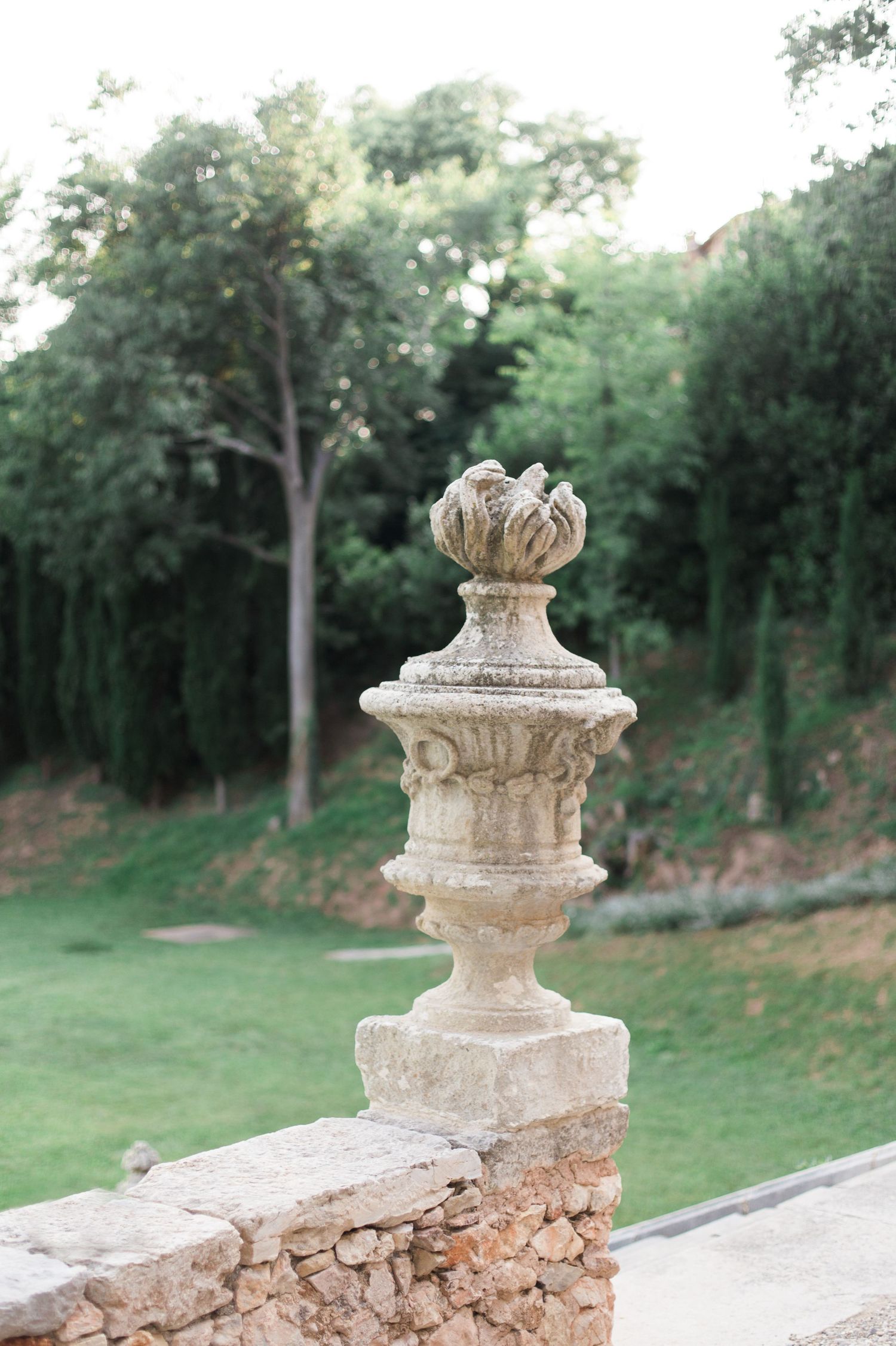 Ornate stone finial decorating a rustic garden wall against lush green foliage.