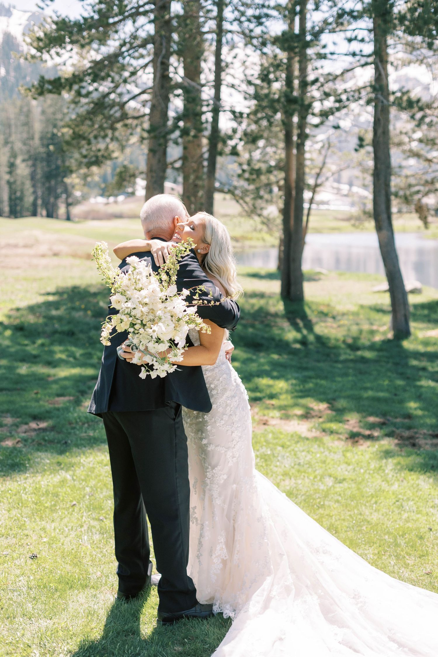 A wedding couple embraces outdoors holding a white floral bouquet against a scenic mountain backdrop.