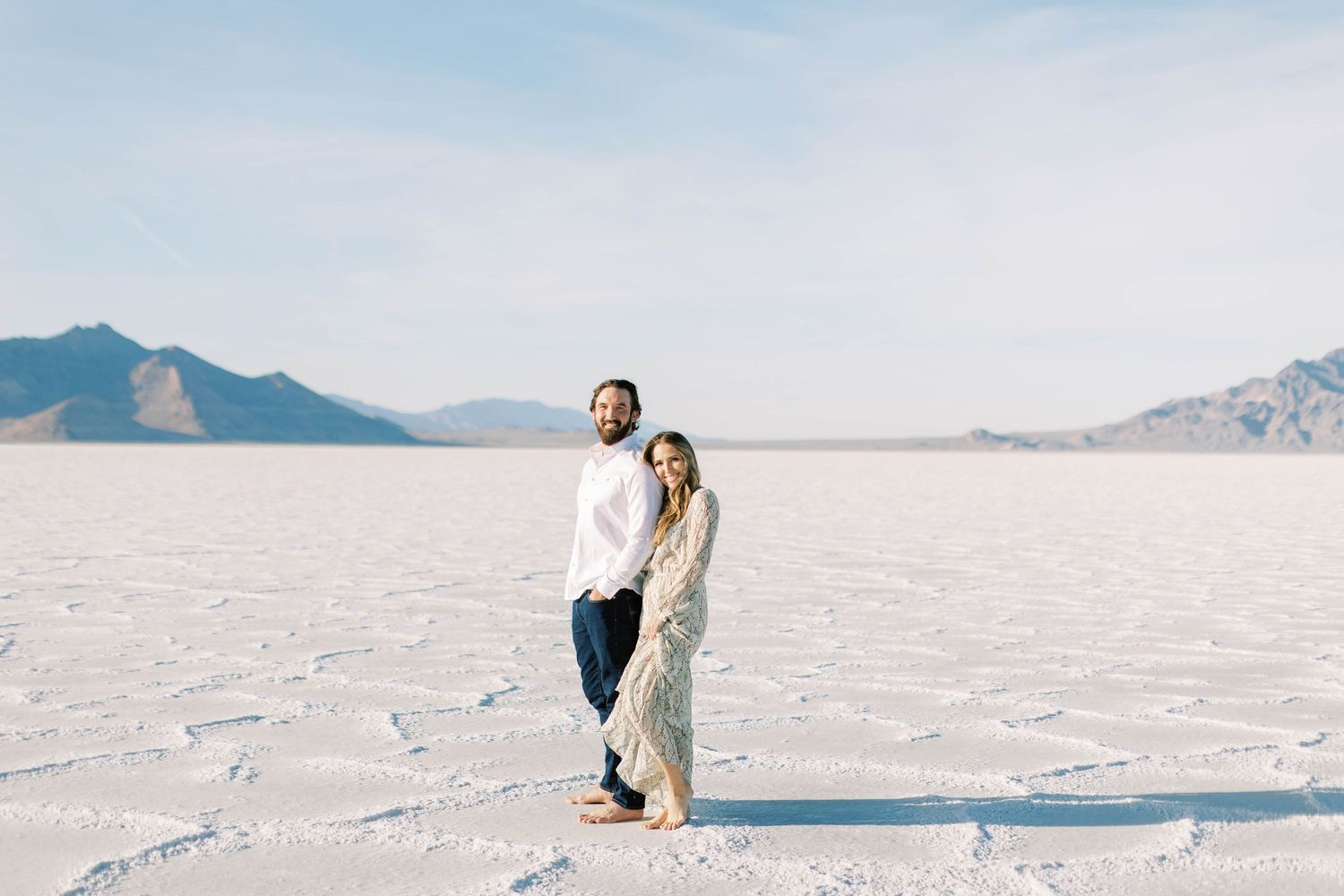 Couple standing together on salt flats with distant mountains under blue sky.