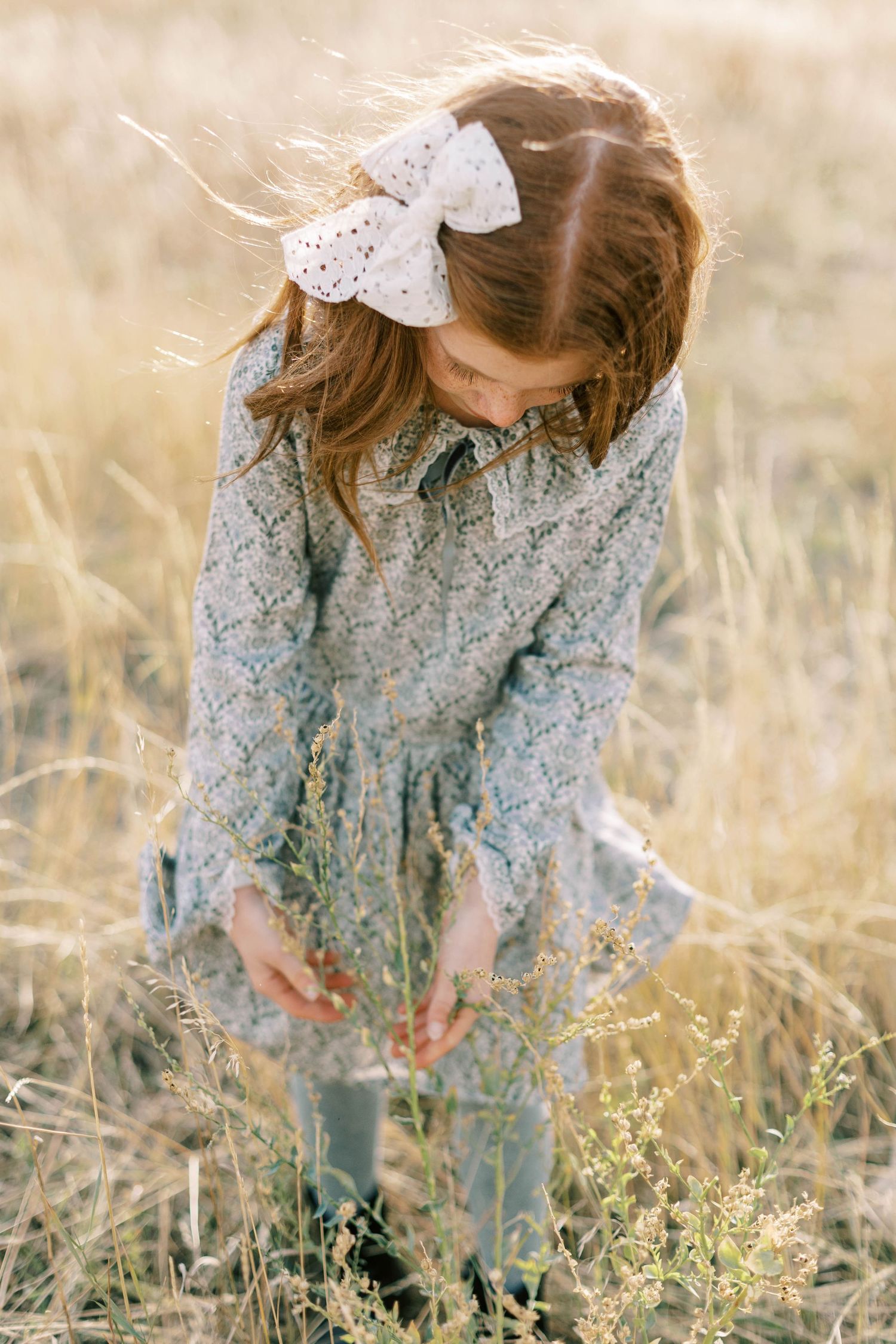 Girl in prairie dress with white bow gathering wildflowers in golden field.