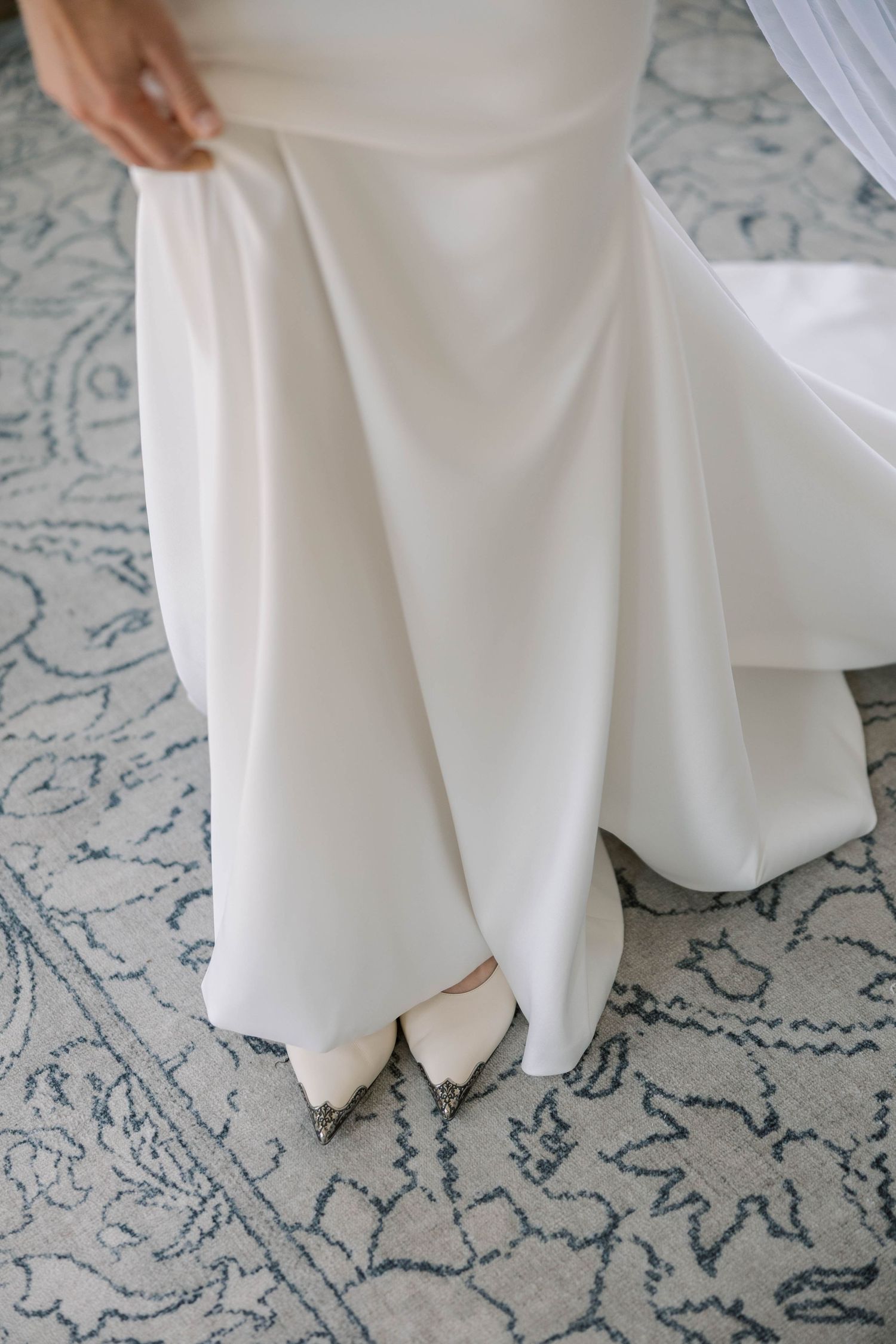 Close-up of flowing white wedding dress hem and shoes against patterned carpet.