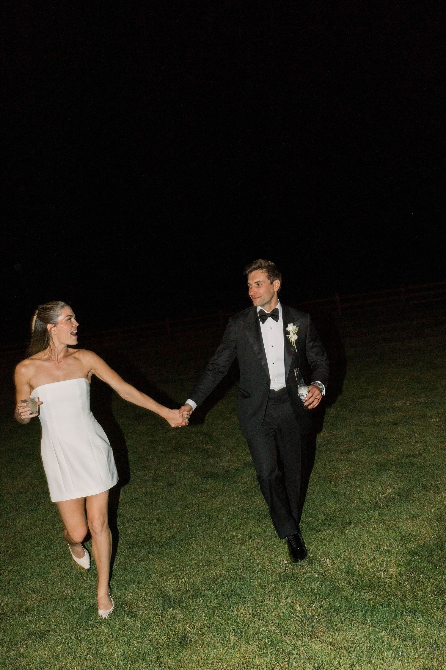 Couple in wedding attire runs together on grass at night holding drinks and hands.