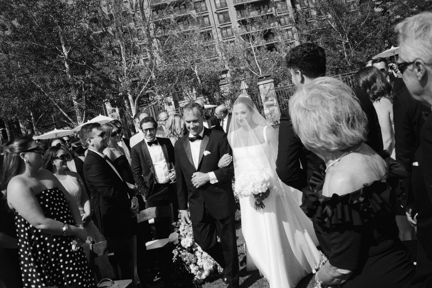 Wedding guests gather outdoors in a natural setting during a black and white photography ceremony celebration.
