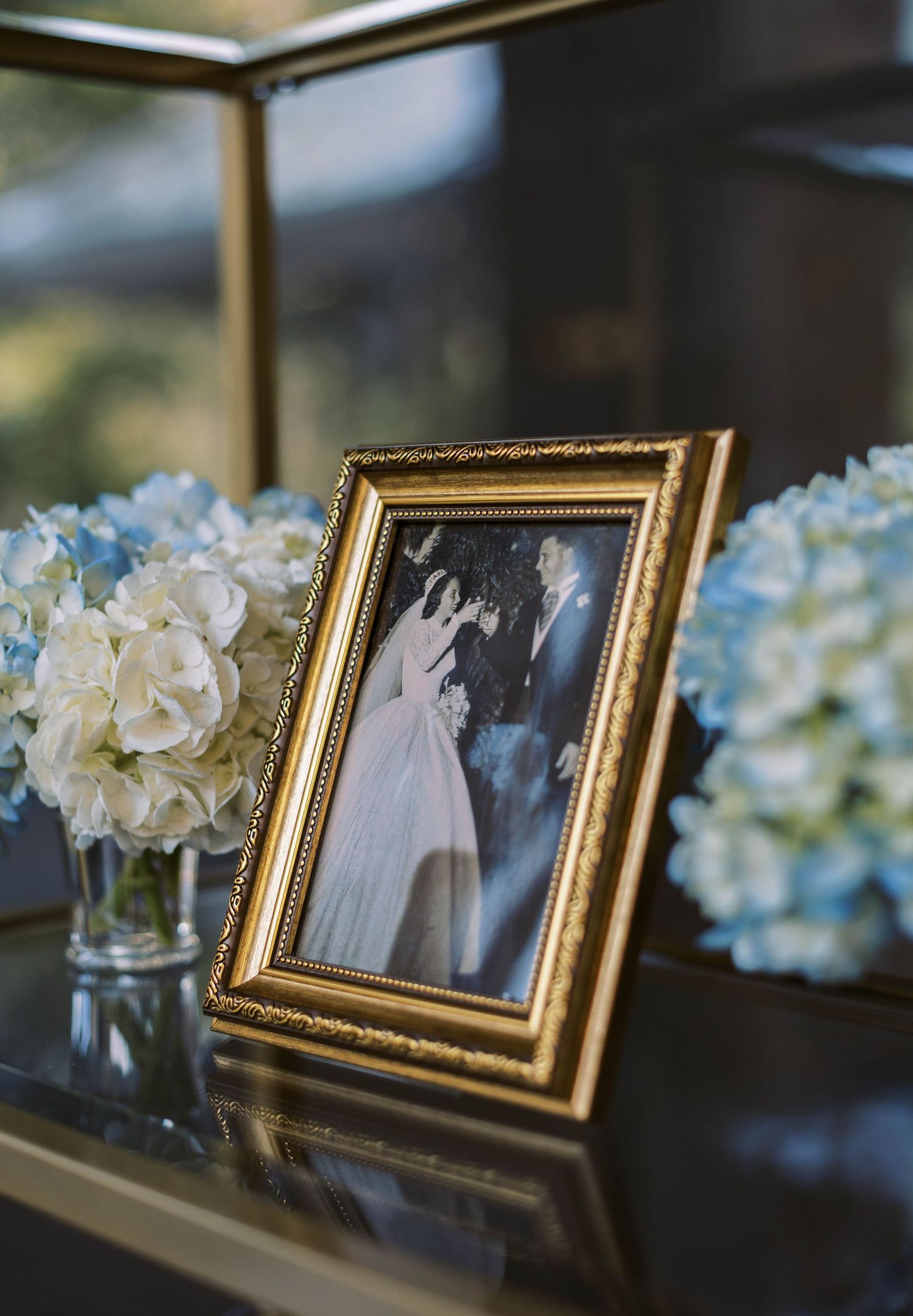 A vintage wedding photo in an ornate gold frame sits on a table with blue and white hydrangea flower arrangements.