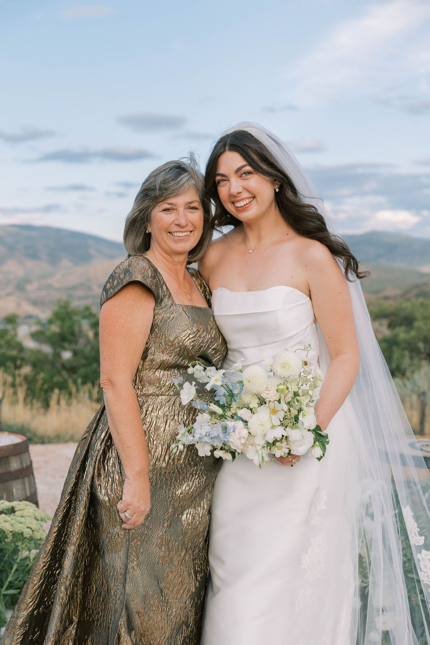 Two women in formal attire share a tender moment outdoors with a mountain landscape in the background.