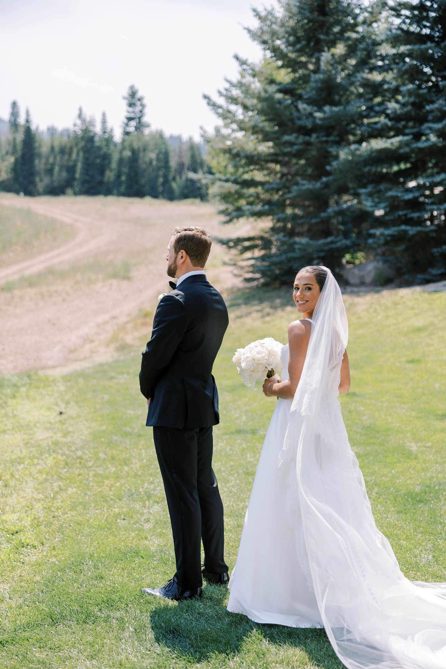 A couple in wedding attire stands on a grassy hillside with evergreen trees and mountains in the background.