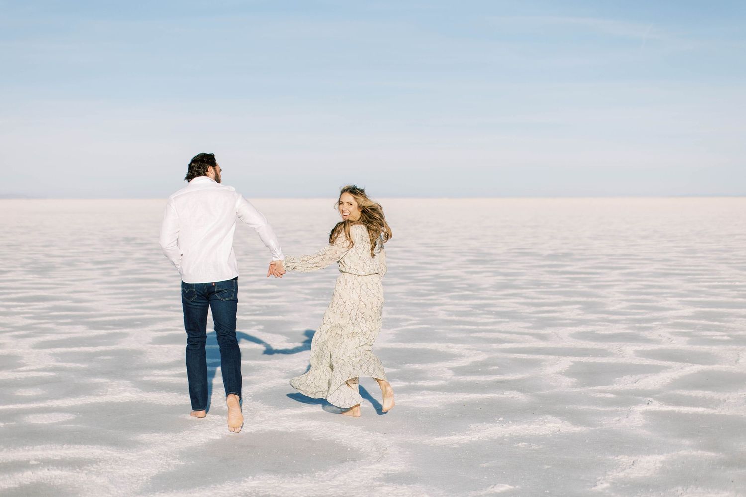 Couple walking hand in hand across white salt flats under bright blue sky.