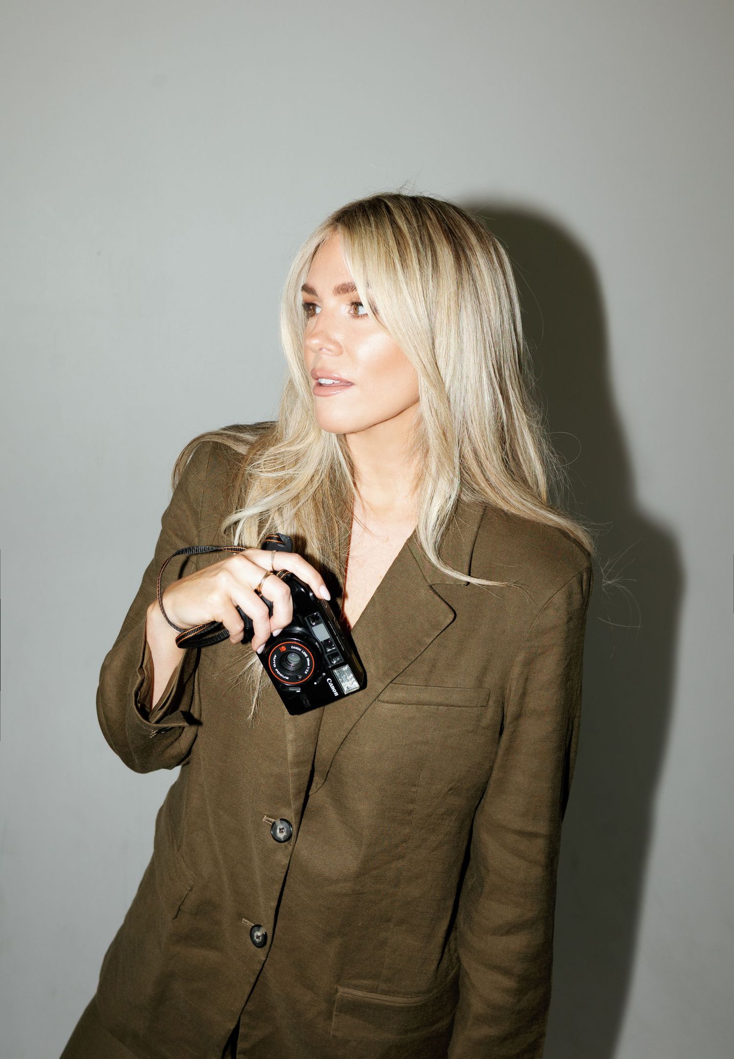 Blonde woman in olive blazer holding a black camera, posing against a gray background.
