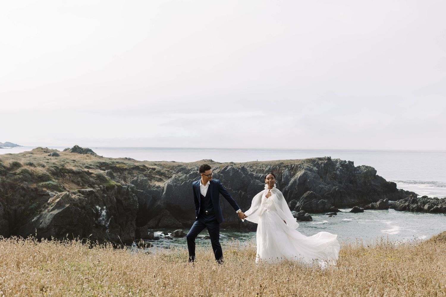 A couple in wedding attire runs through a grassy coastal field overlooking dramatic ocean cliffs on a cloudy day.