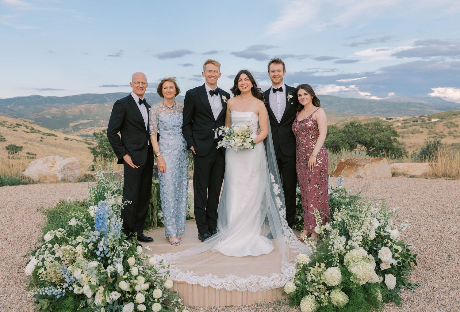 Wedding party poses together outdoors against a scenic desert mountain backdrop with white floral arrangements.