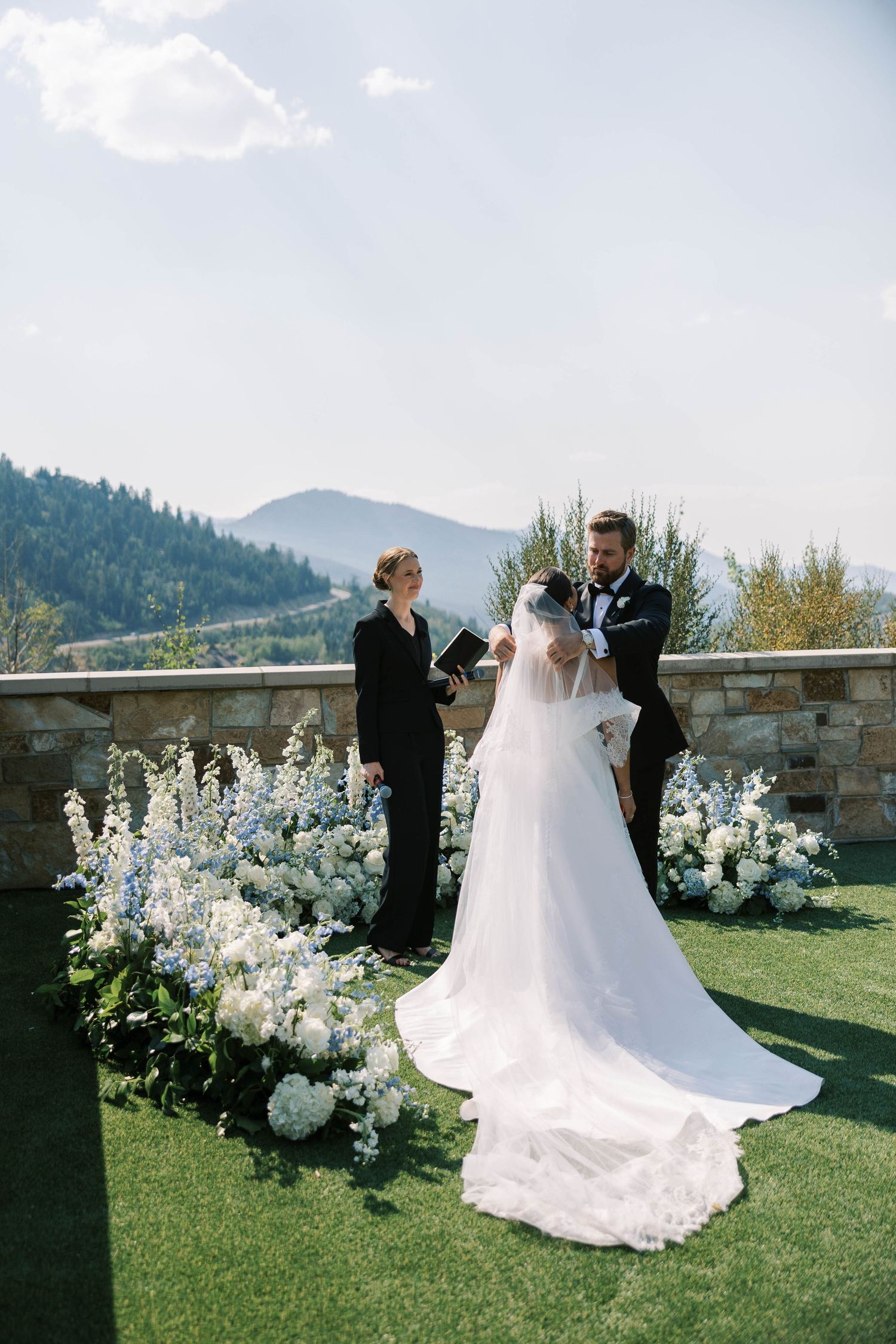 A wedding ceremony takes place on a scenic mountain overlook with white floral arrangements and a stone wall backdrop.