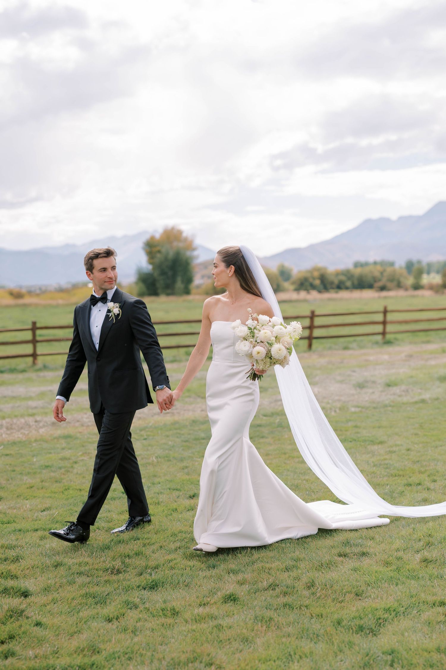 Newlyweds walk hand in hand across a grassy field with mountains and wooden fences in the background.
