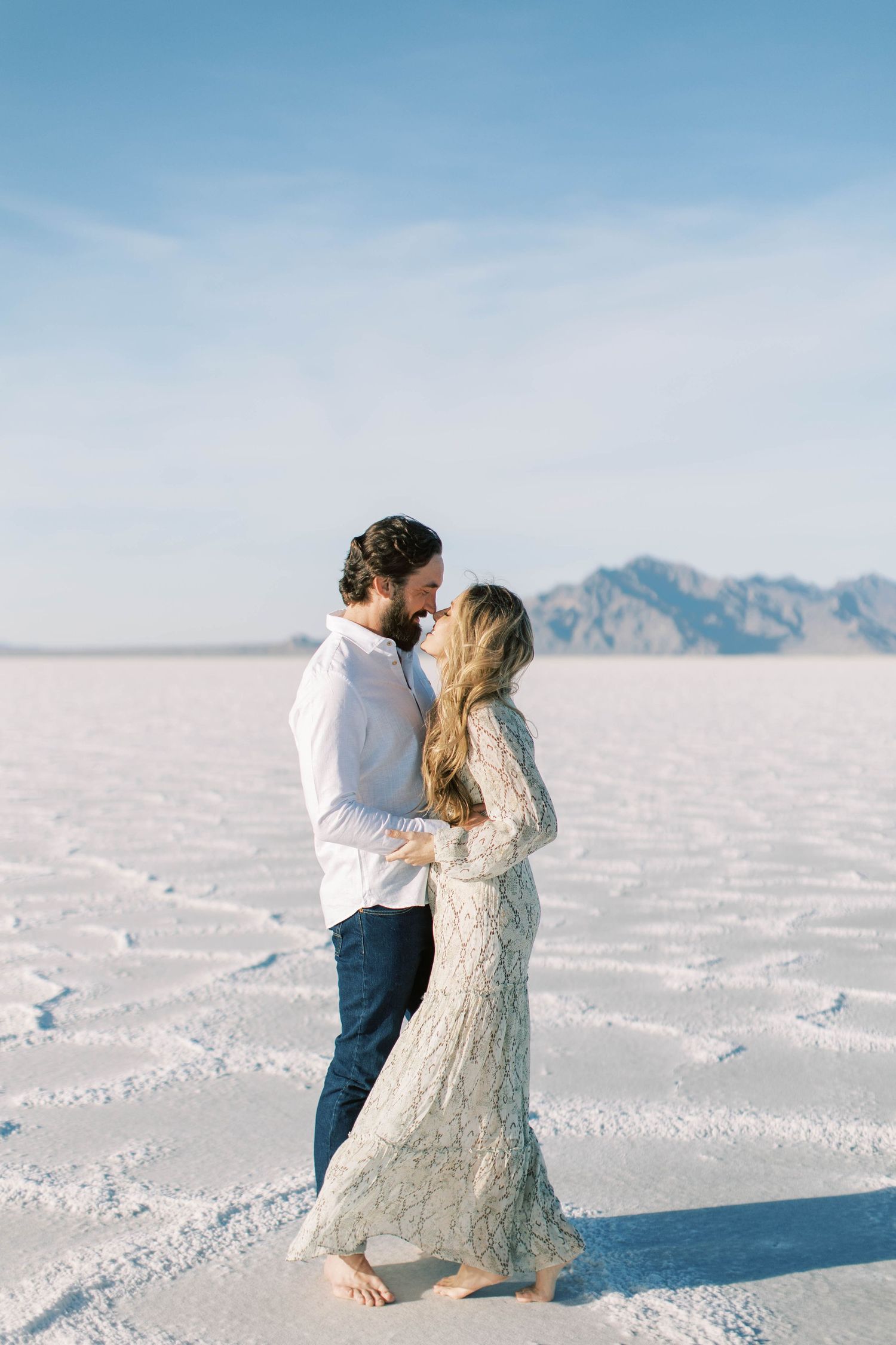 Couple in white attire dancing on salt flats with mountains in background.