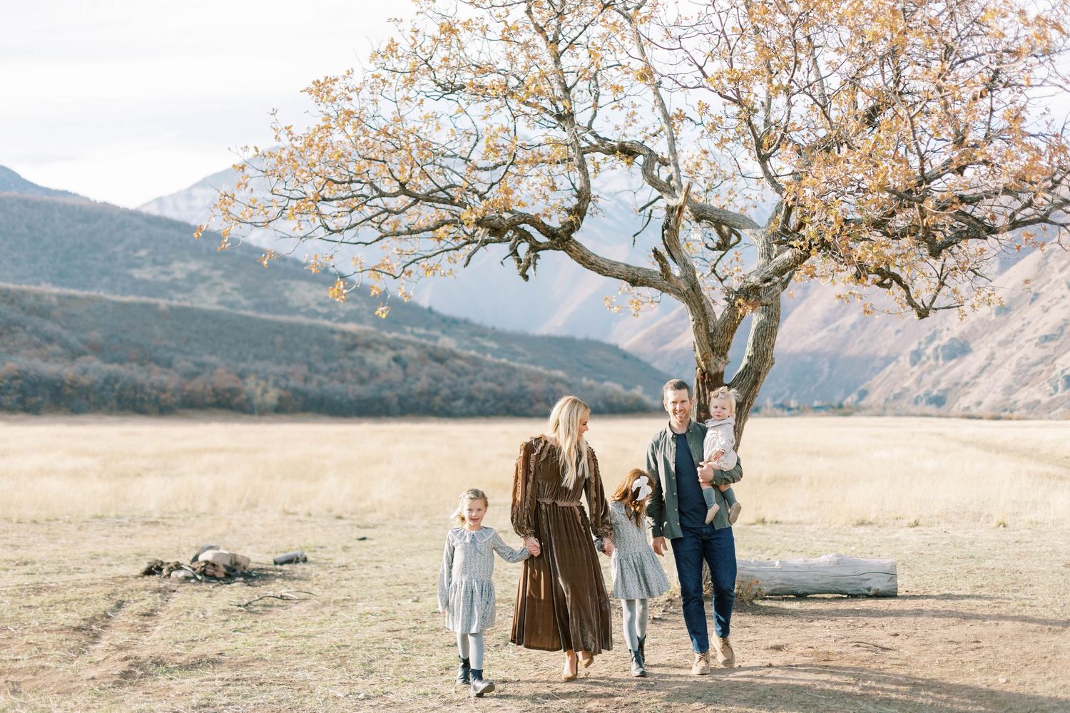 A family stands together under a bare tree in a scenic mountain valley during autumn.