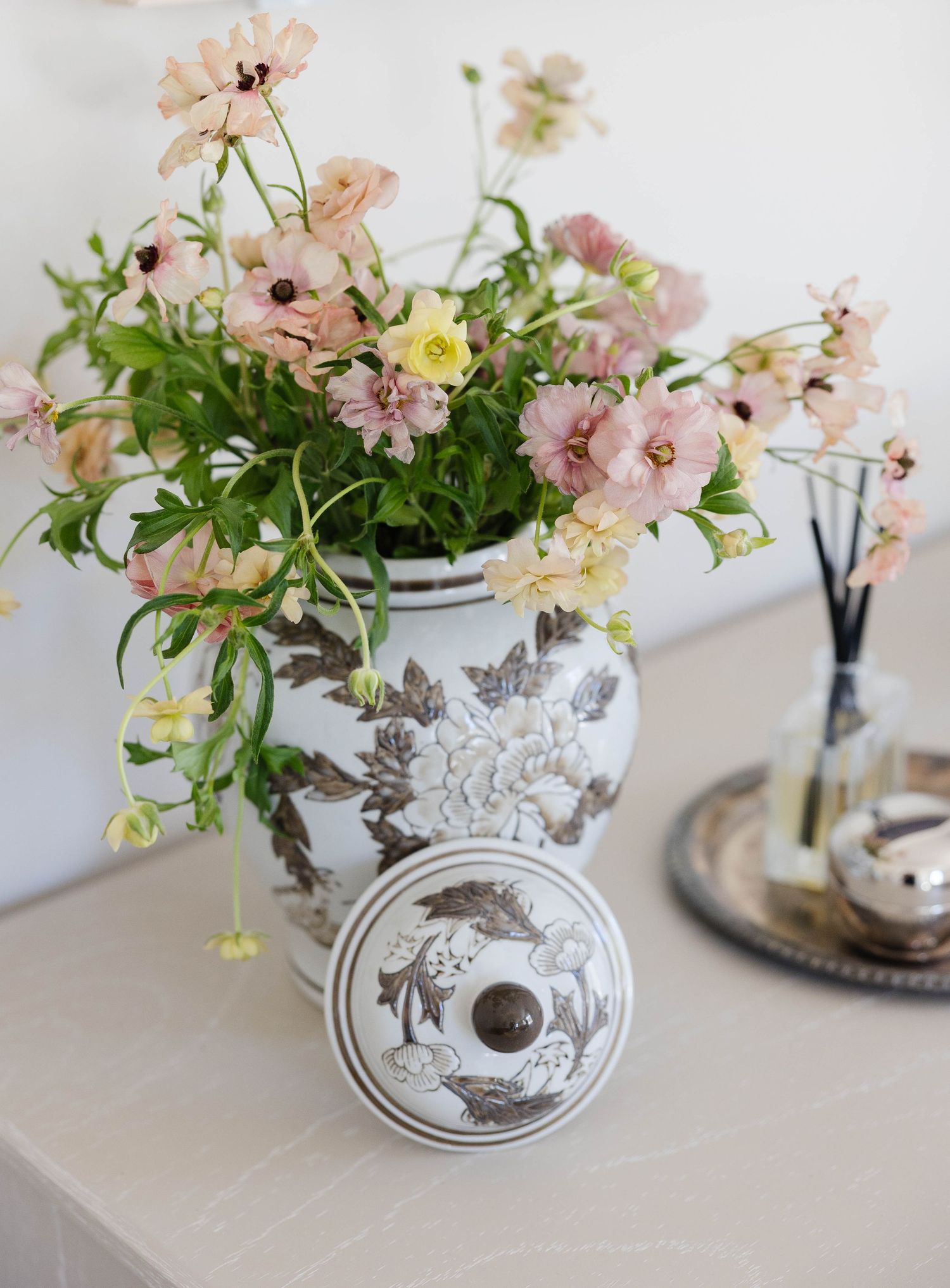 Pink cosmos flowers arranged in a white ceramic ginger jar with brown floral motifs on a marble surface.