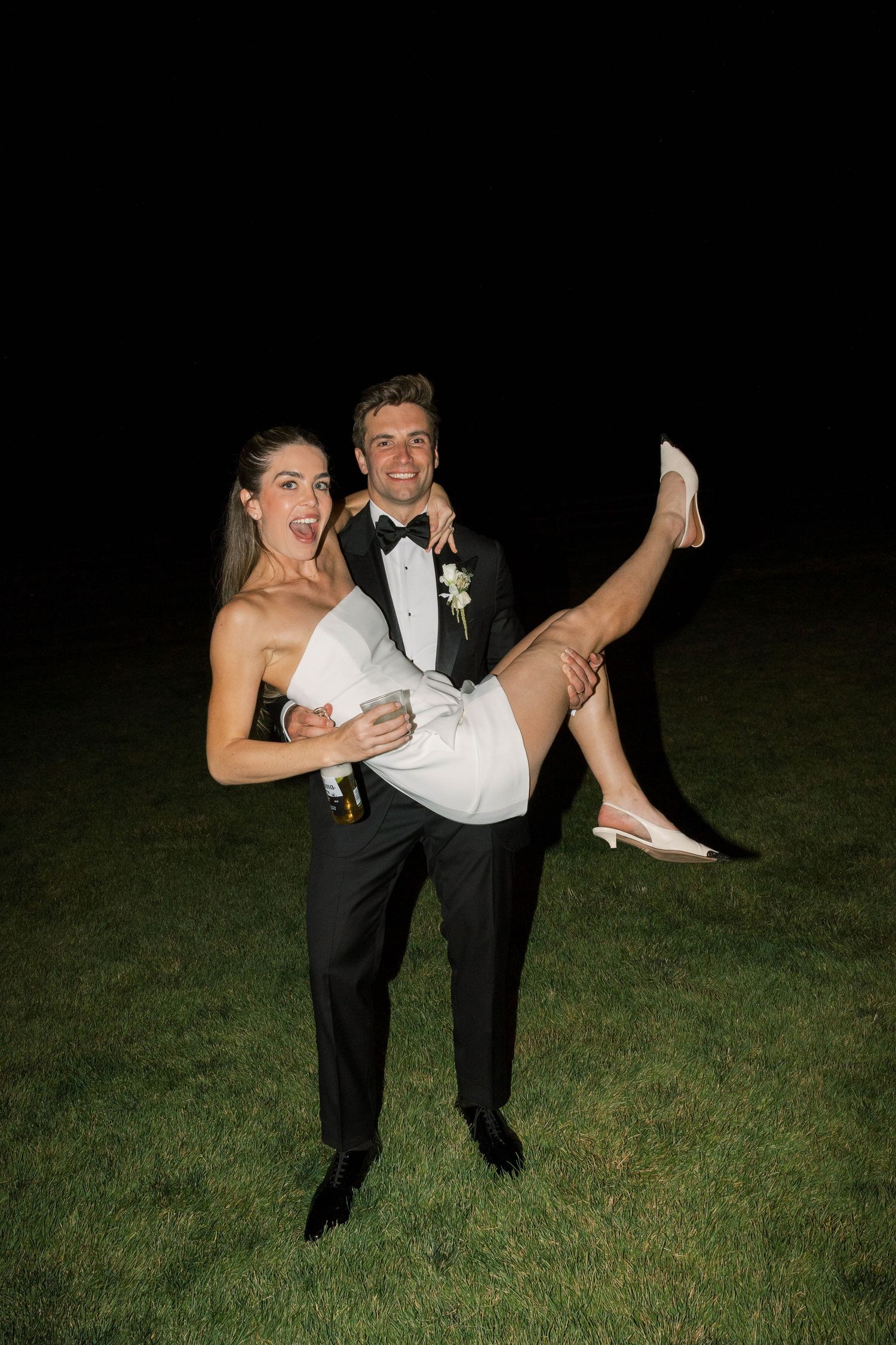 A couple in formal wedding attire playfully pose on a dark grassy lawn at night.
