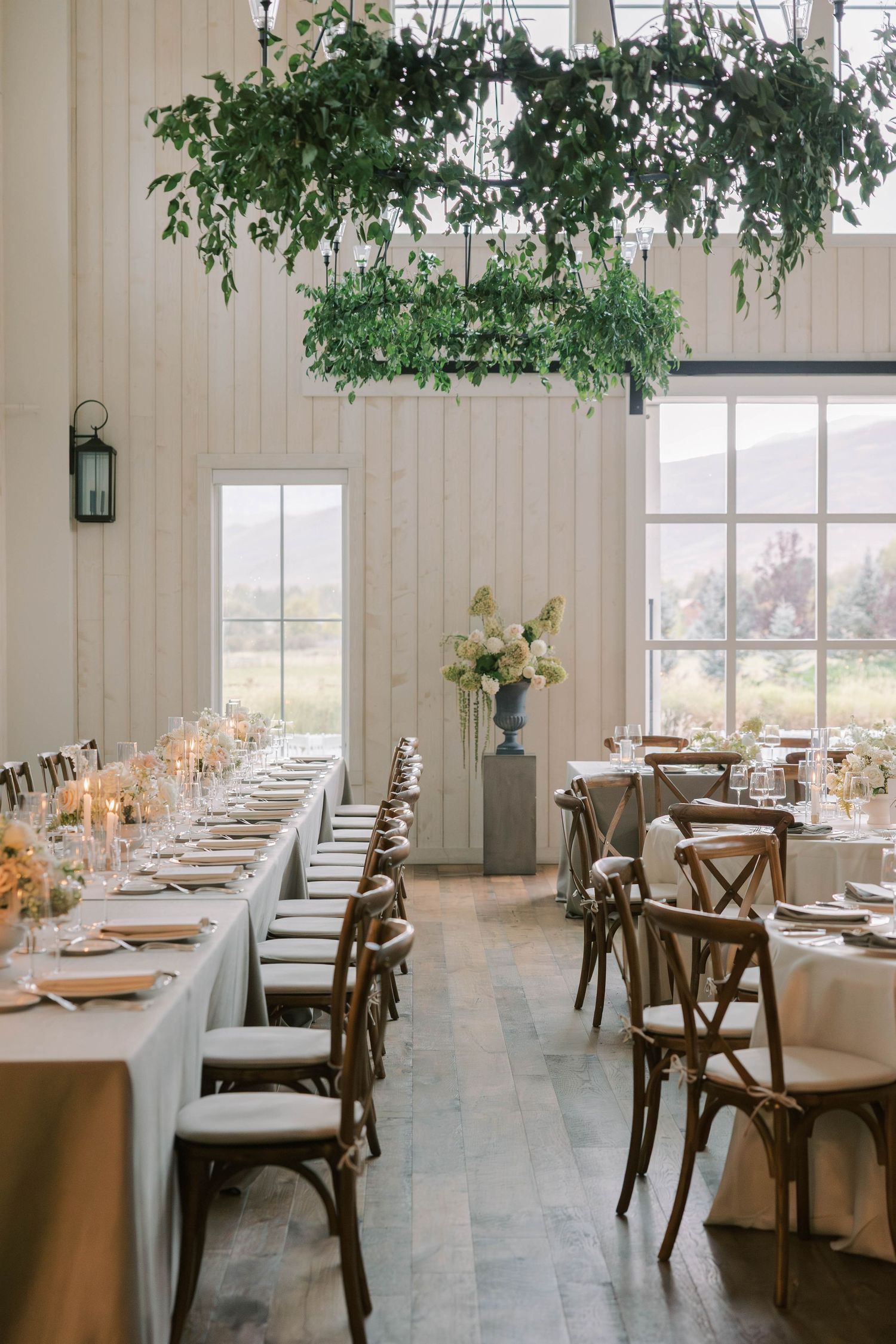 Elegant wedding reception setup with long white-clothed tables and wooden crossback chairs in a bright venue with large windows.