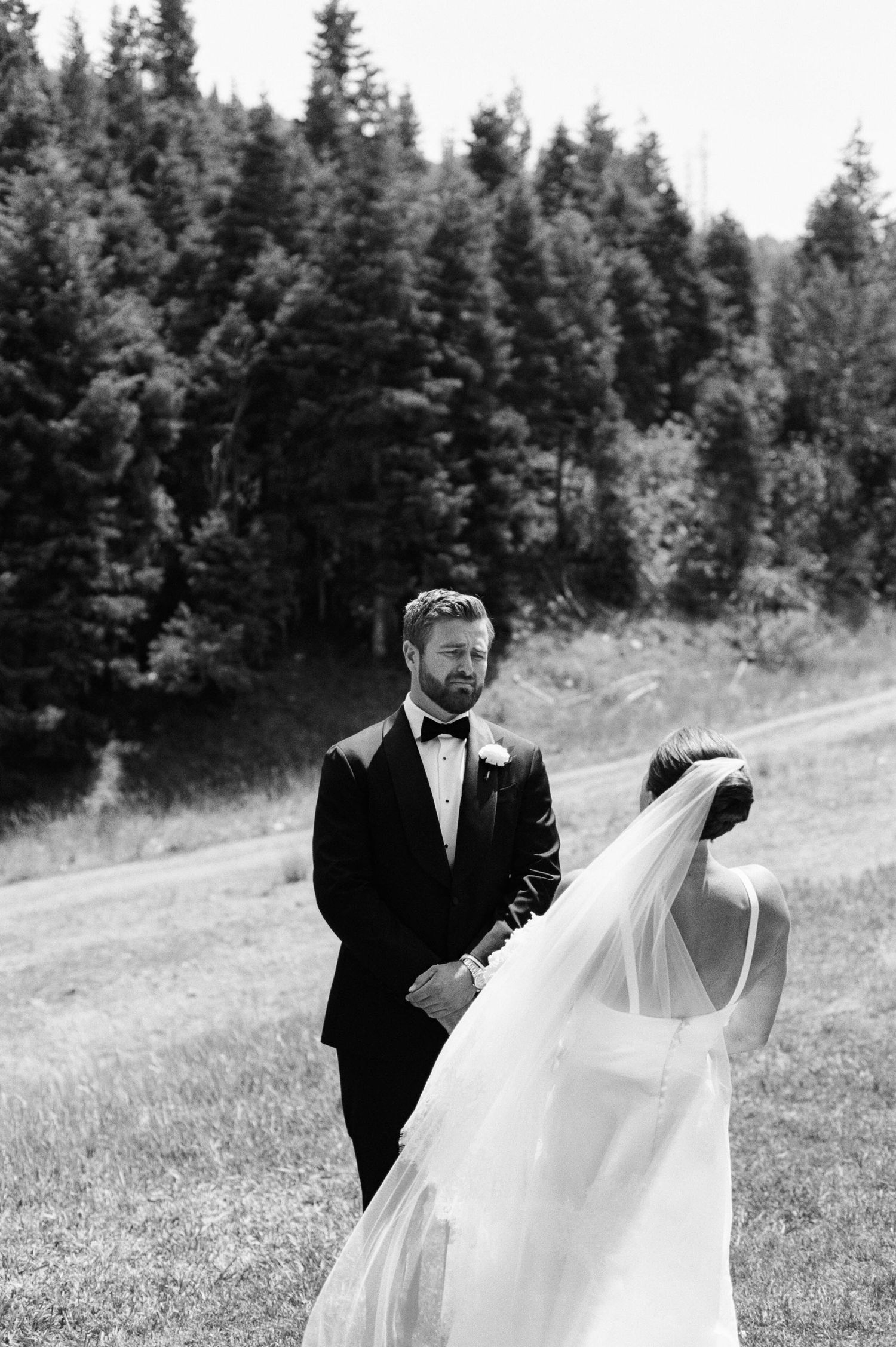 A couple shares an intimate moment in a meadow with evergreen trees in the background, captured in black and white.
