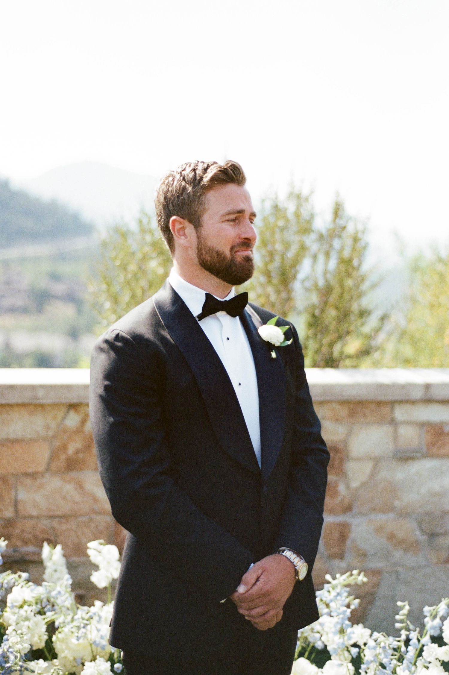 Elegant groom in black tuxedo and bowtie stands outdoors by stone wall with white floral arrangements at scenic venue.