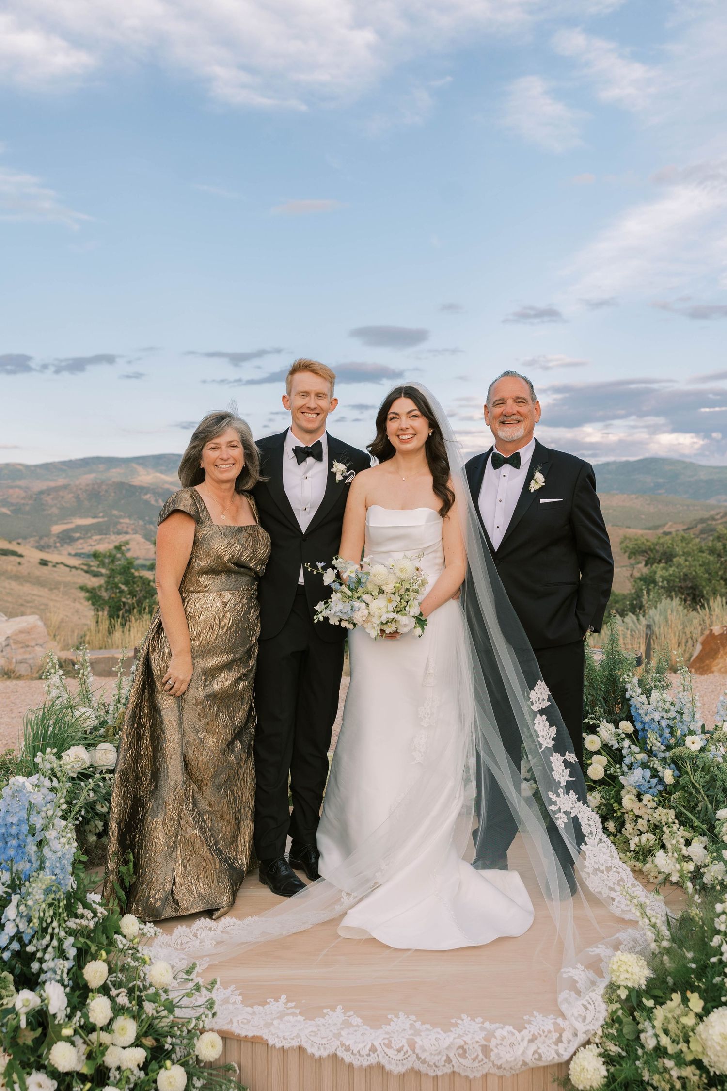 Wedding party poses outdoors against mountain backdrop with floral arch and elegant decor.
