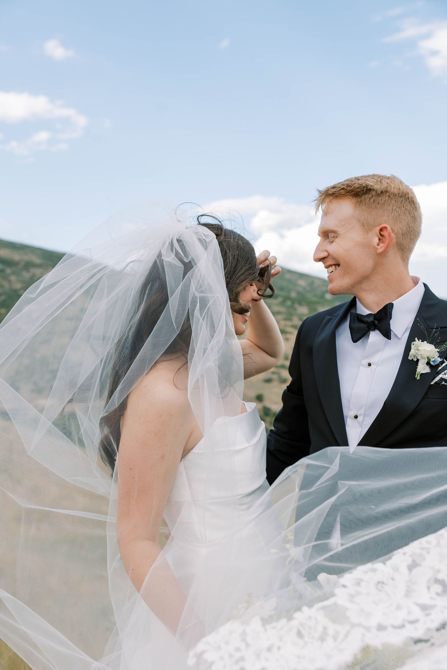 Wedding couple embracing outdoors with bride's veil flowing dramatically in mountain setting.