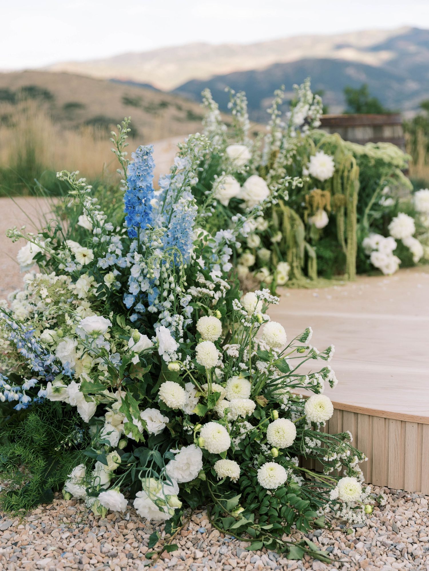 Elegant floral arrangement with white roses and blue delphiniums against mountain backdrop.