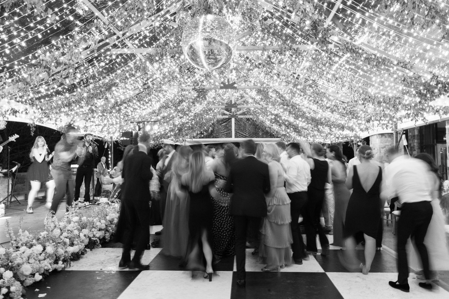 Black and white panoramic photo shows a large group of people dancing under decorative lights at an indoor event.