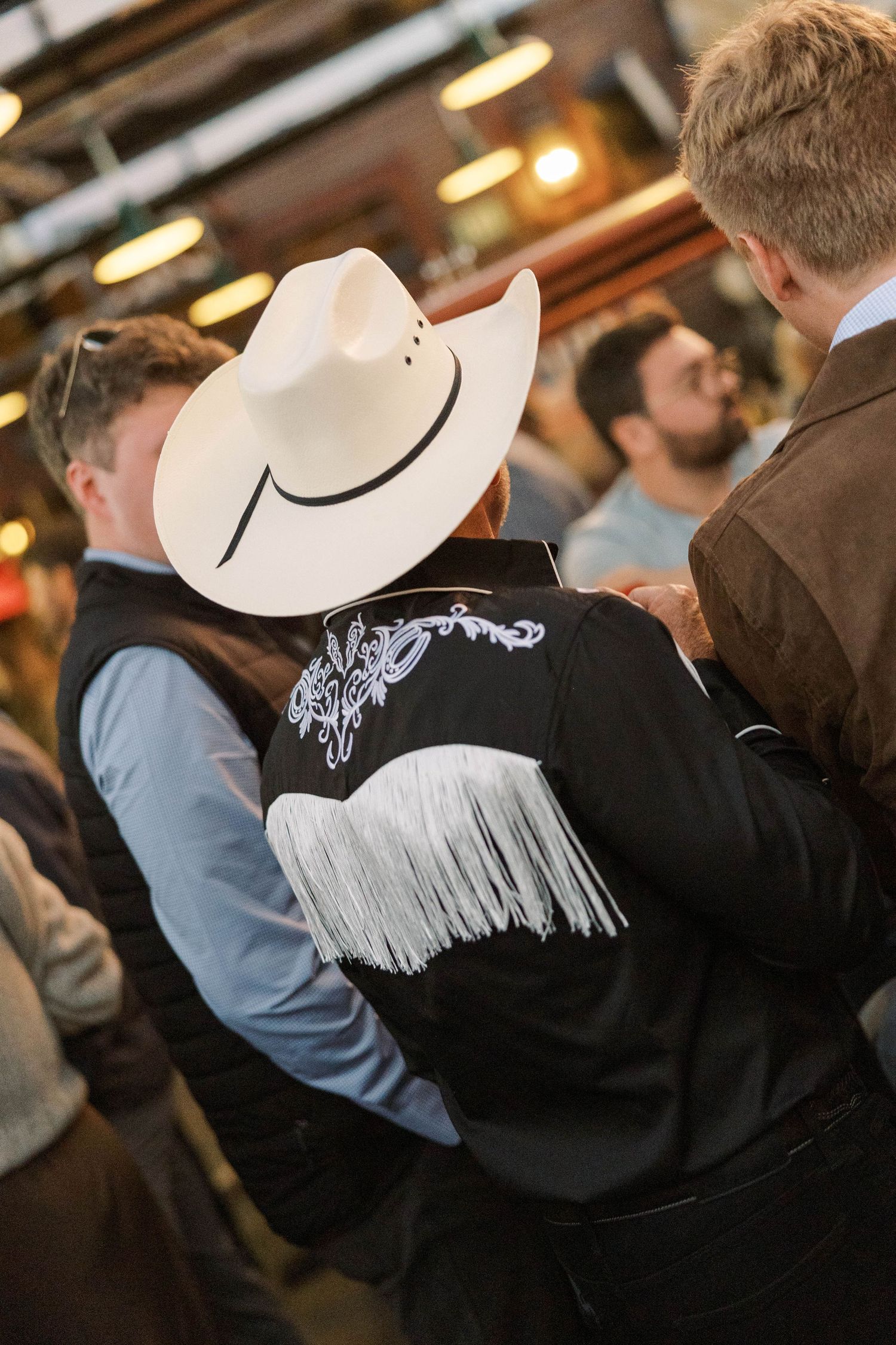 Person wearing a white cowboy hat and black jacket with embroidered design in a crowded indoor setting.