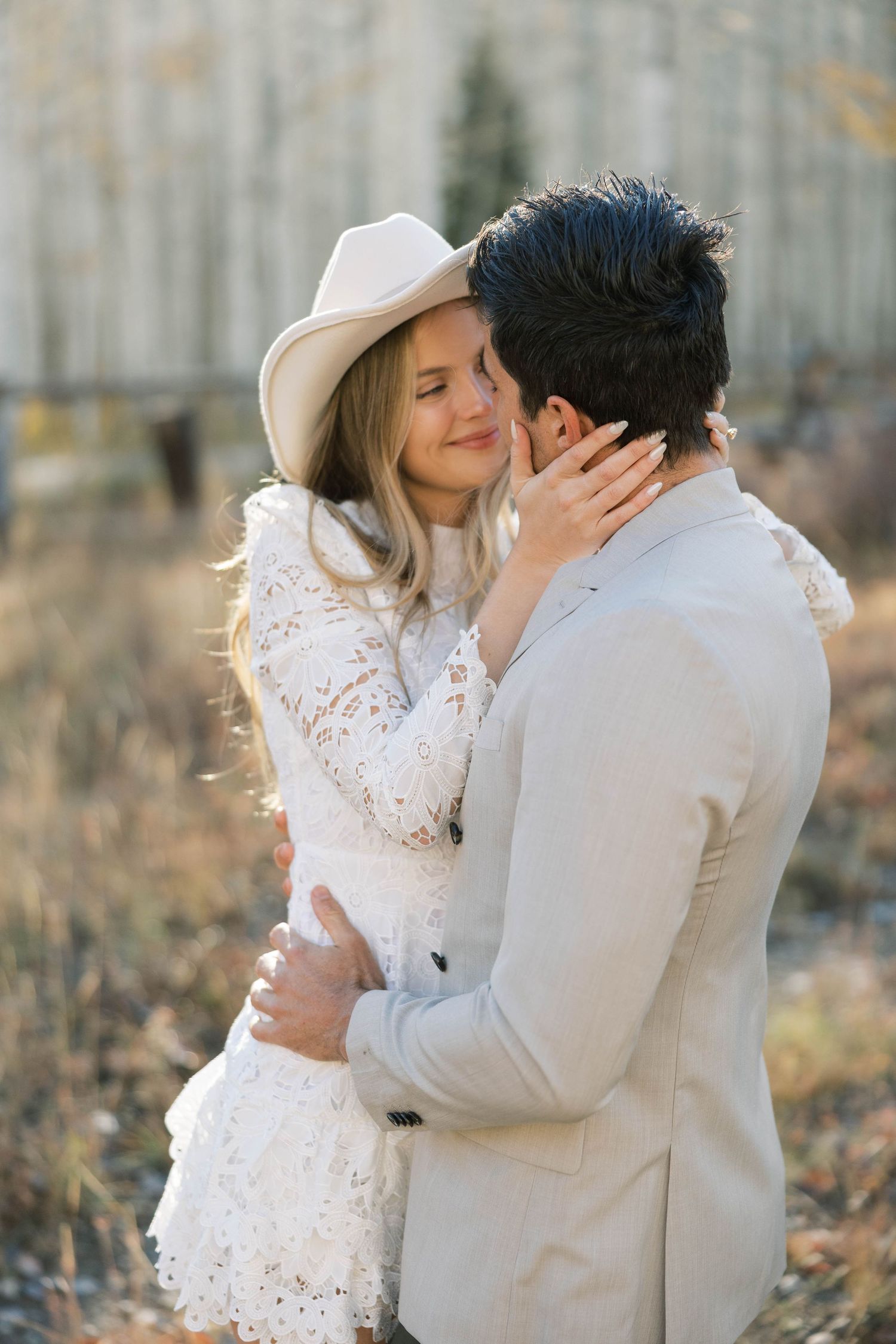 Romantic couple embraces outdoors in autumn, with woman in white lace dress and cowboy hat sharing tender moment.