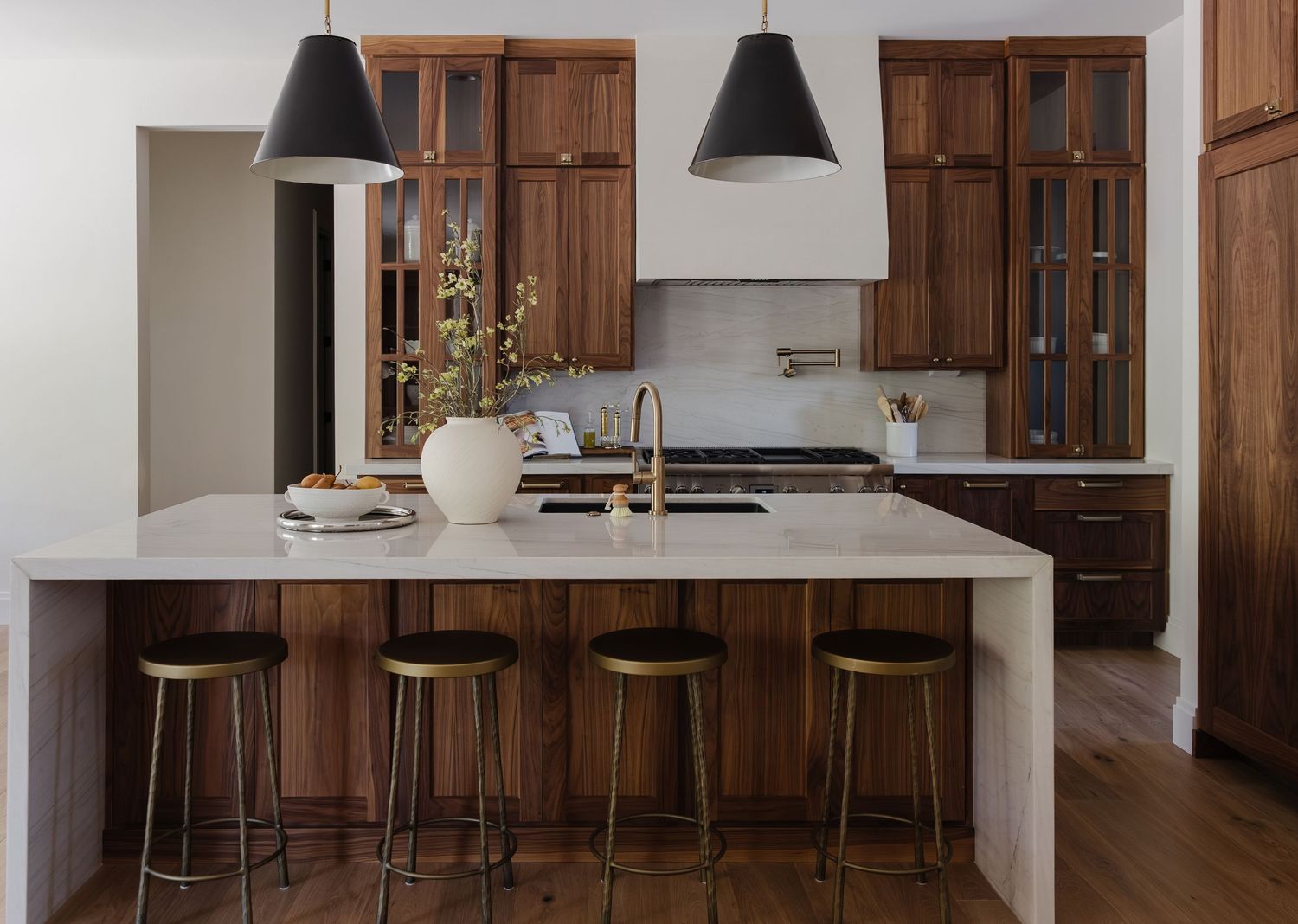 Modern kitchen with walnut cabinets, marble island, gold bar stools, brass faucet, and black pendant lights.