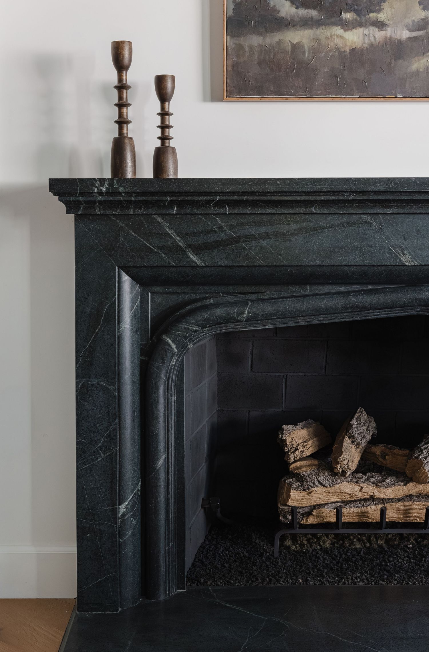 Dark green marble fireplace mantel with logs inside, two wooden candlesticks on top, and a landscape painting above.