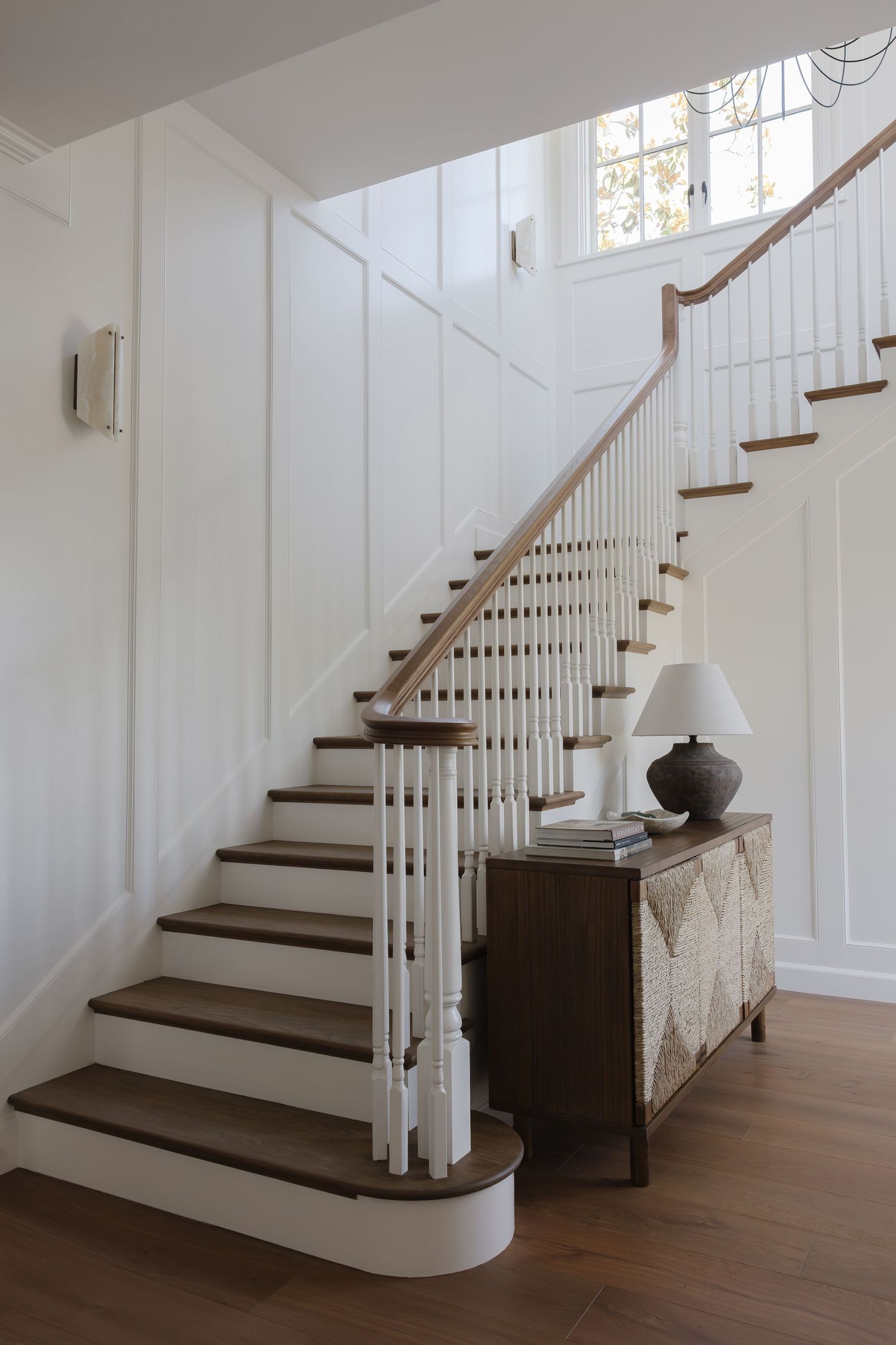Elegant staircase with white balusters, dark wood treads, and a vintage console table with lamp in a bright white foyer.