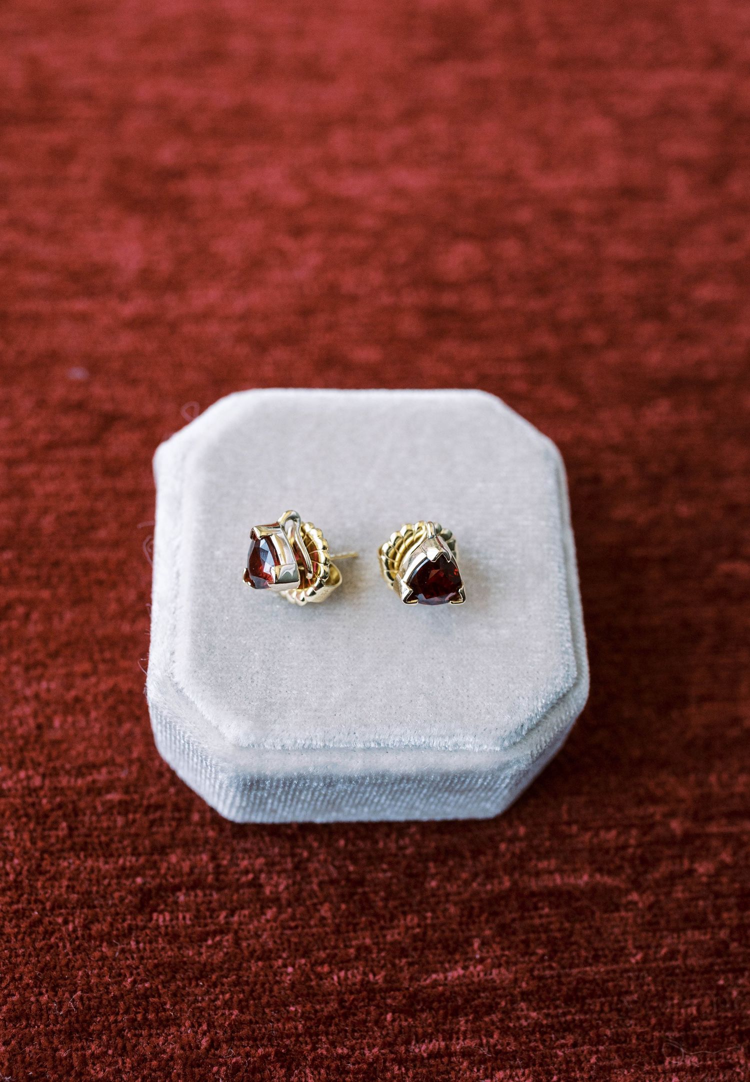 A pair of delicate gold stud earrings with dark stones displayed in a light gray velvet ring box on burgundy fabric.