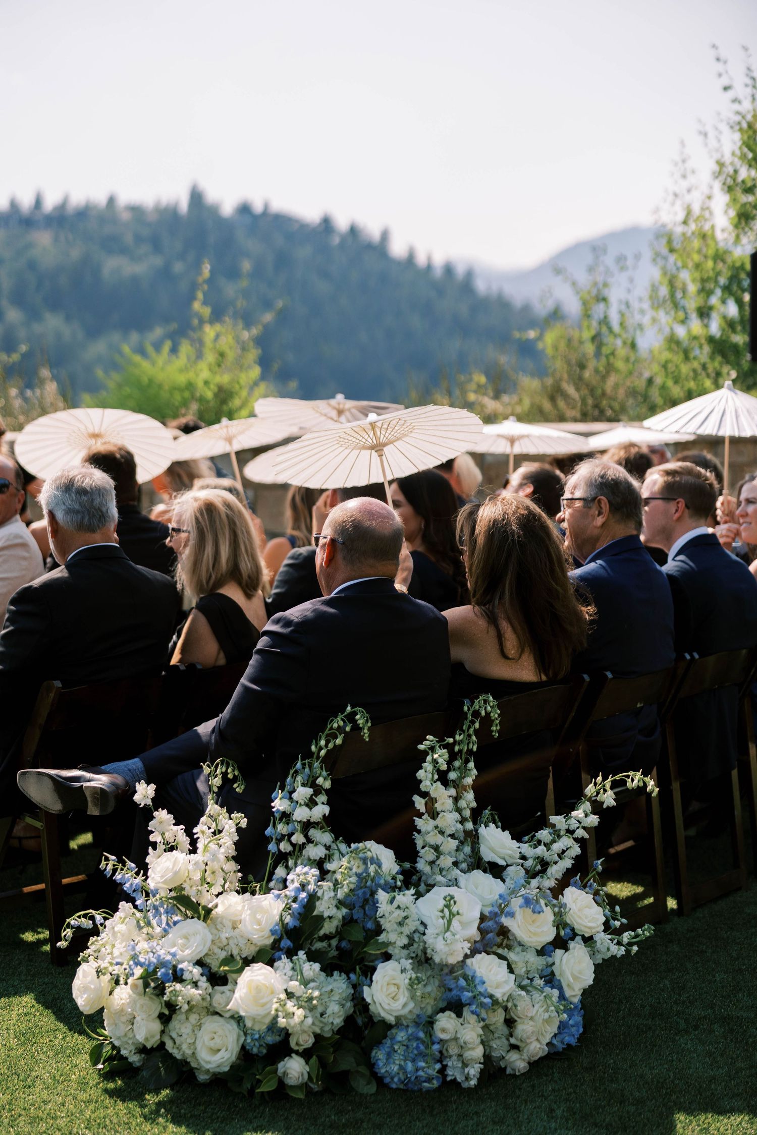 Wedding guests sit outdoors with white parasols and floral arrangements against a mountain backdrop during a ceremony.