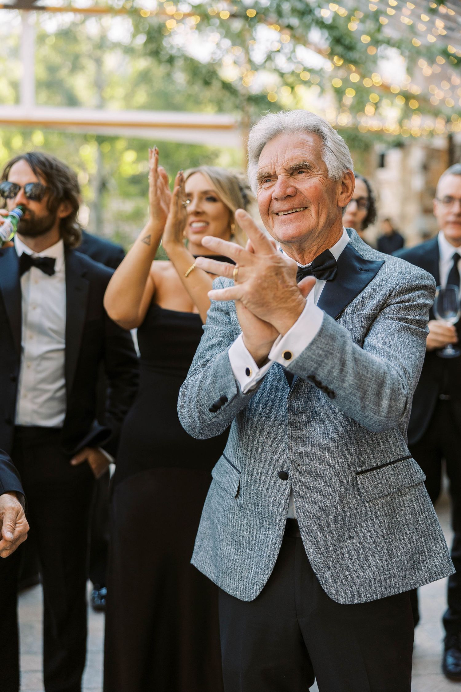 Wedding guests in formal attire clap and celebrate during an outdoor reception under string lights.