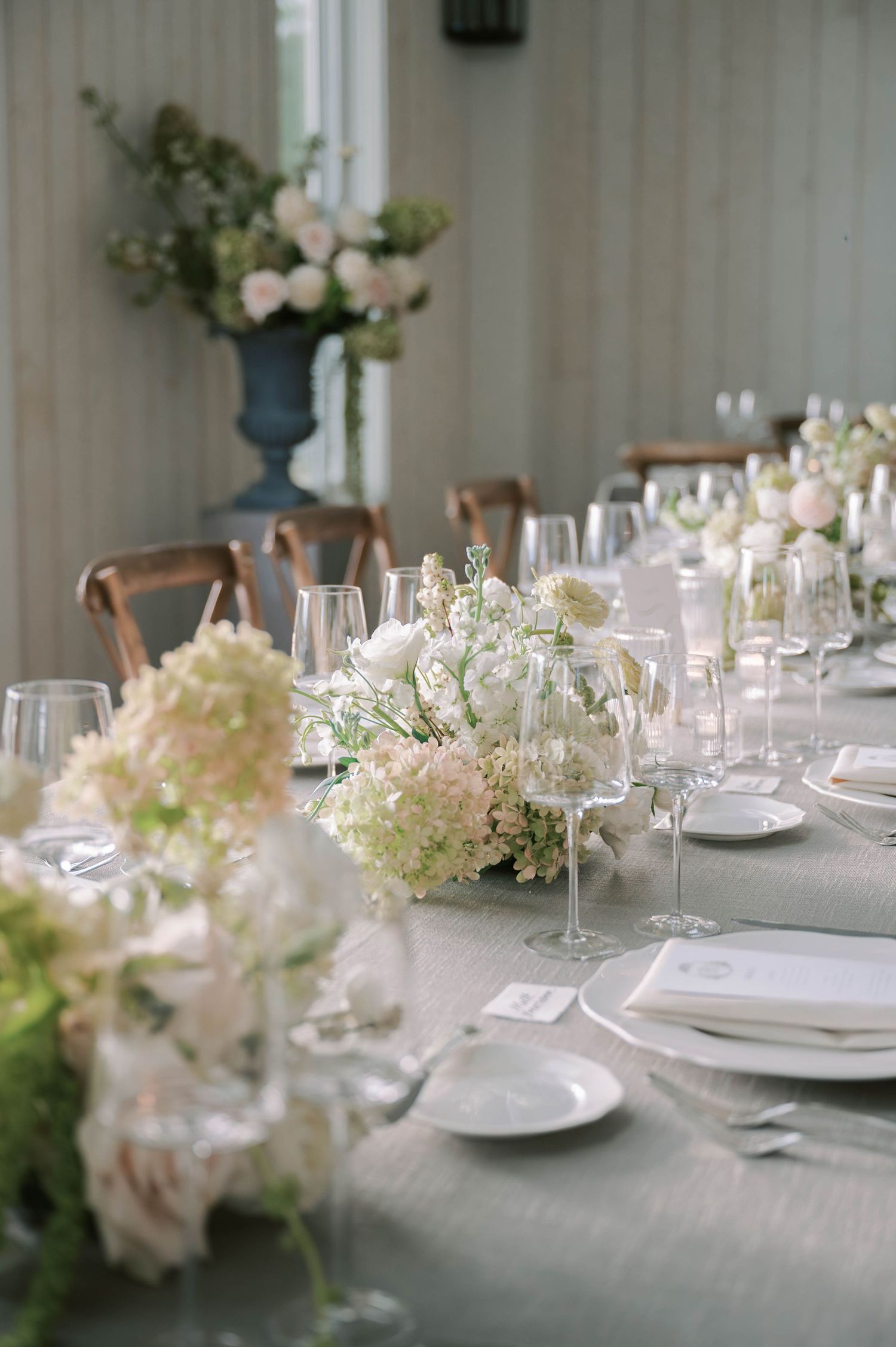 Close-up of formal table settings with white plates, wine glasses, and soft pink floral centerpieces on a neutral tablecloth.
