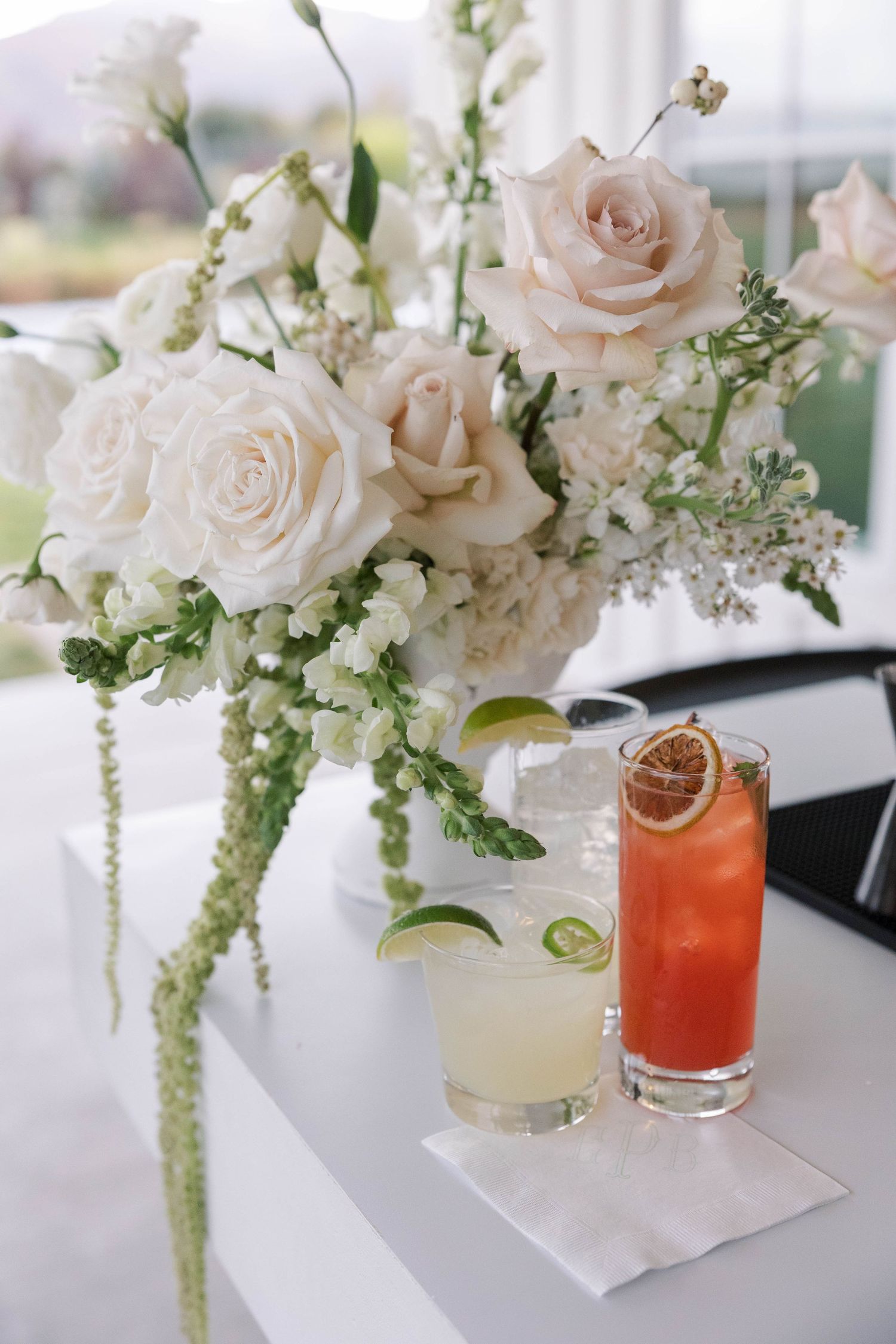 Elegant floral centerpieces with blush roses and white flowers alongside colorful cocktails on a white tablecloth.