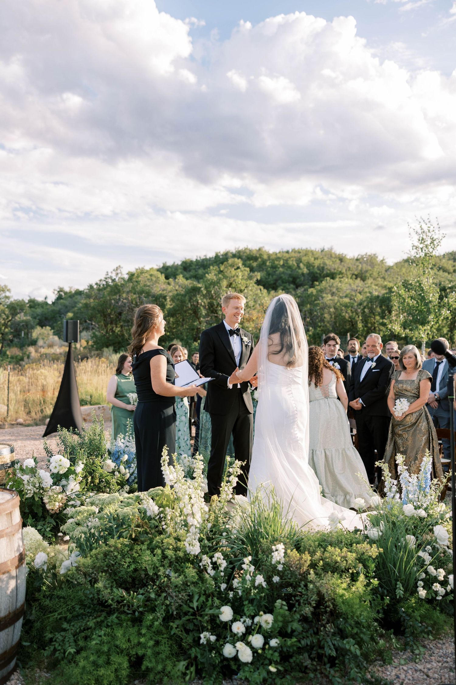 Outdoor wedding ceremony with guests gathered among white floral arrangements in mountain setting.