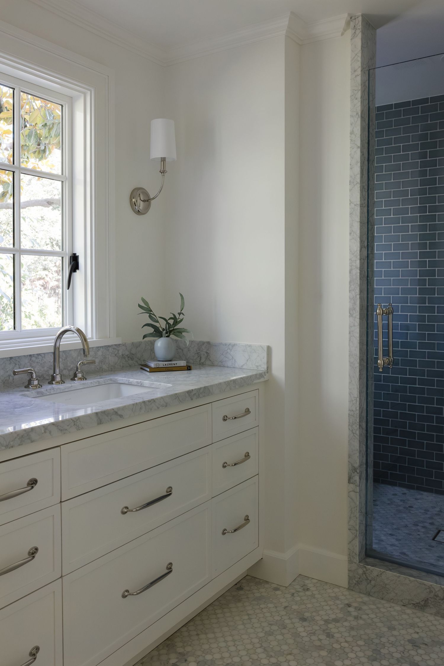 White bathroom vanity with marble countertop, wall sconce, and blue subway tile walk-in shower with brass fixtures.
