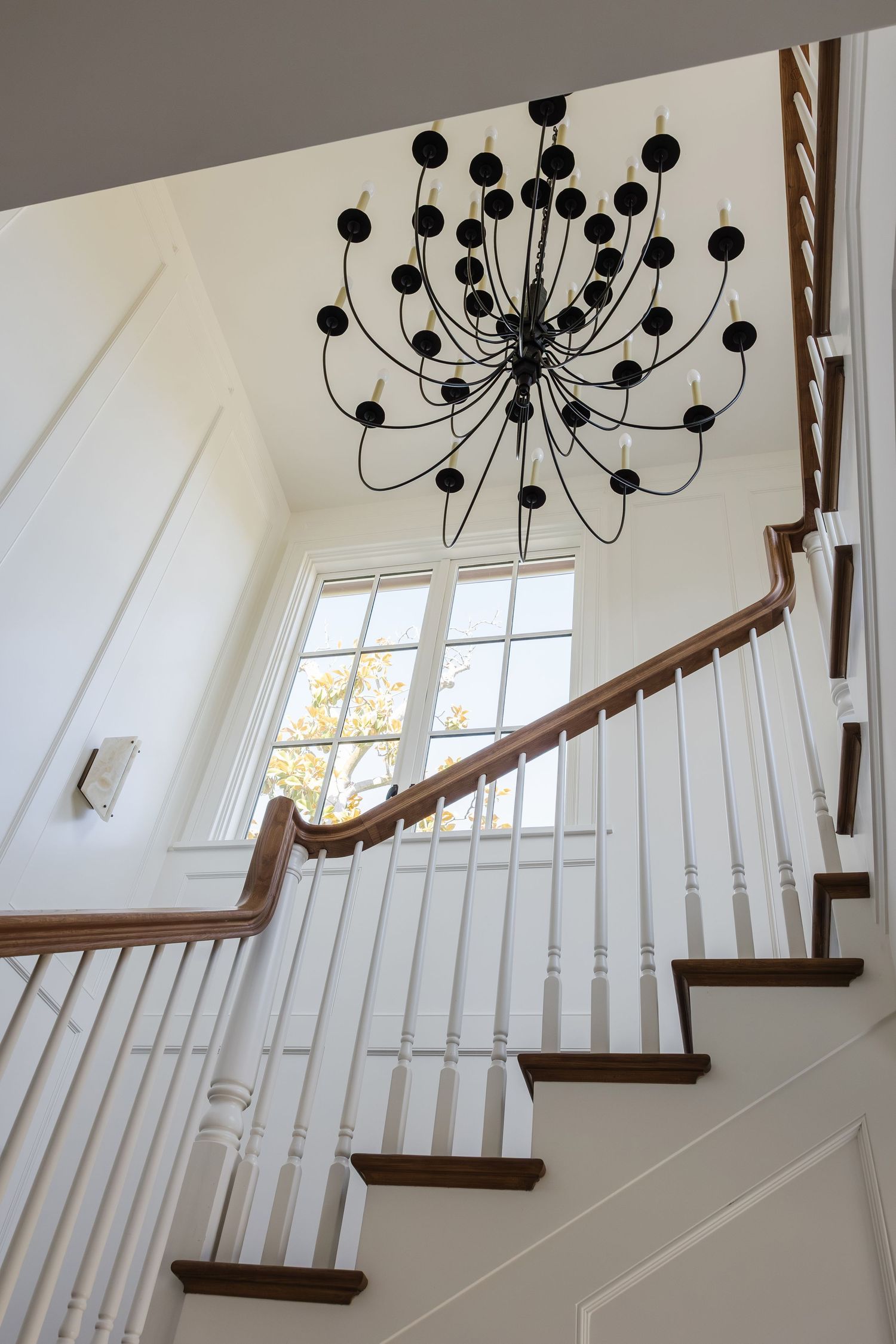 Elegant foyer staircase with white balusters, wood handrail, and a black modern chandelier against bright white walls.