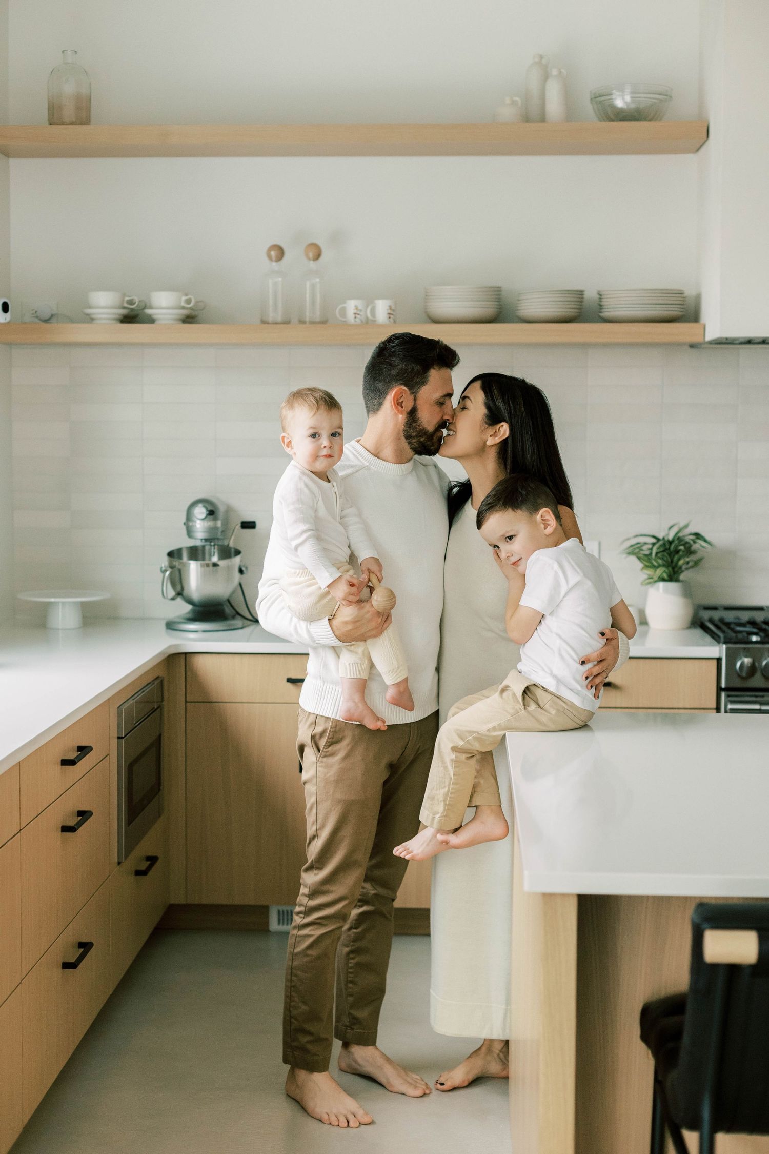 Sweet family moment in modern kitchen with wood cabinets and floating shelves.