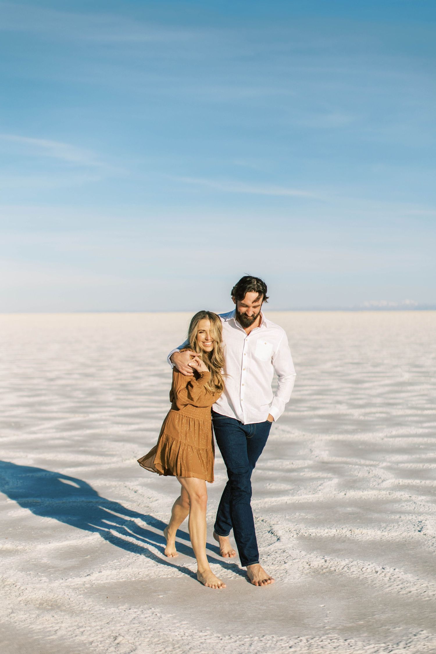 Romantic couple embracing while standing barefoot on white salt flats with mountains in distance.