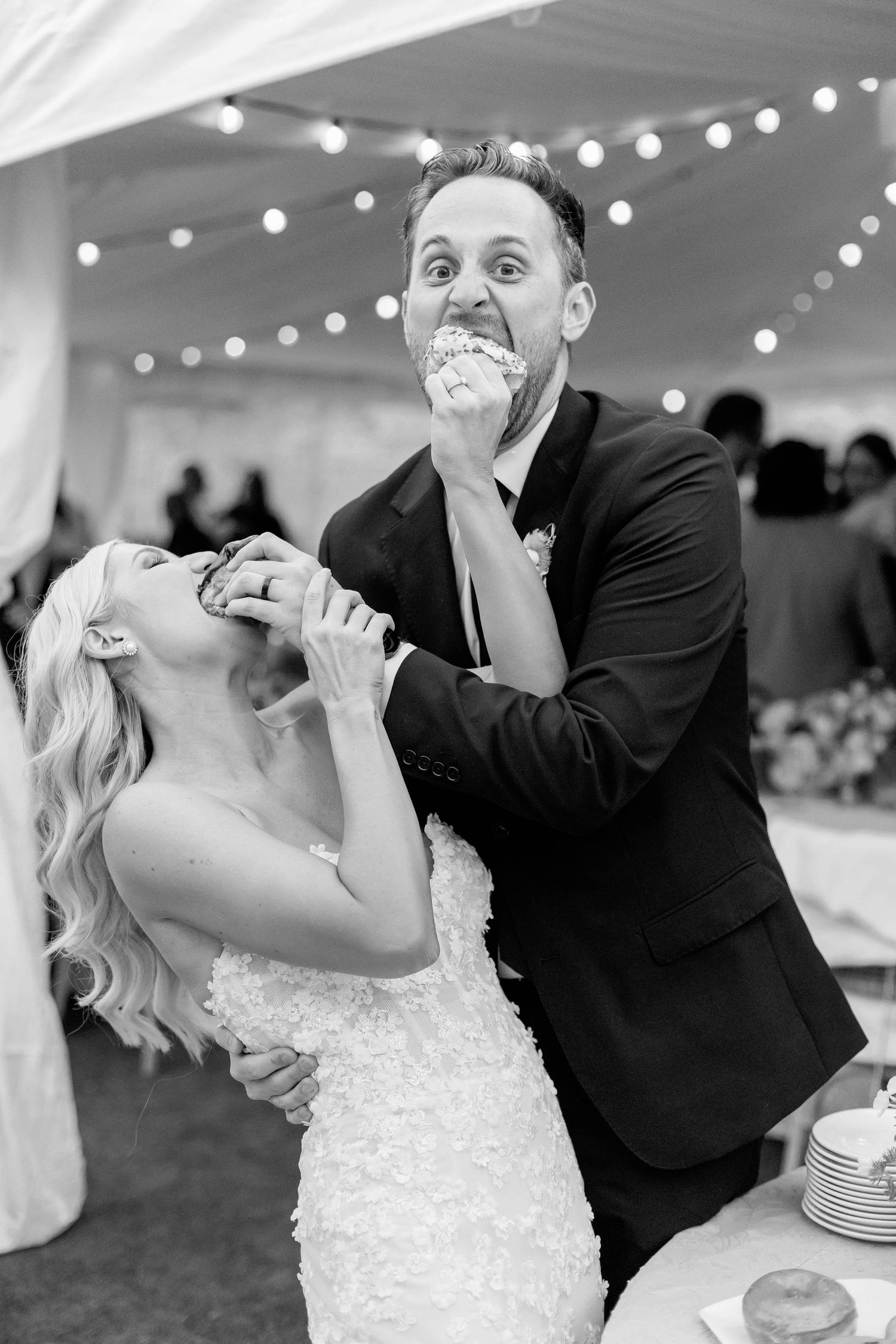 A playful moment captured at a wedding reception as newlyweds laugh while sharing cake under string lights in a tent.