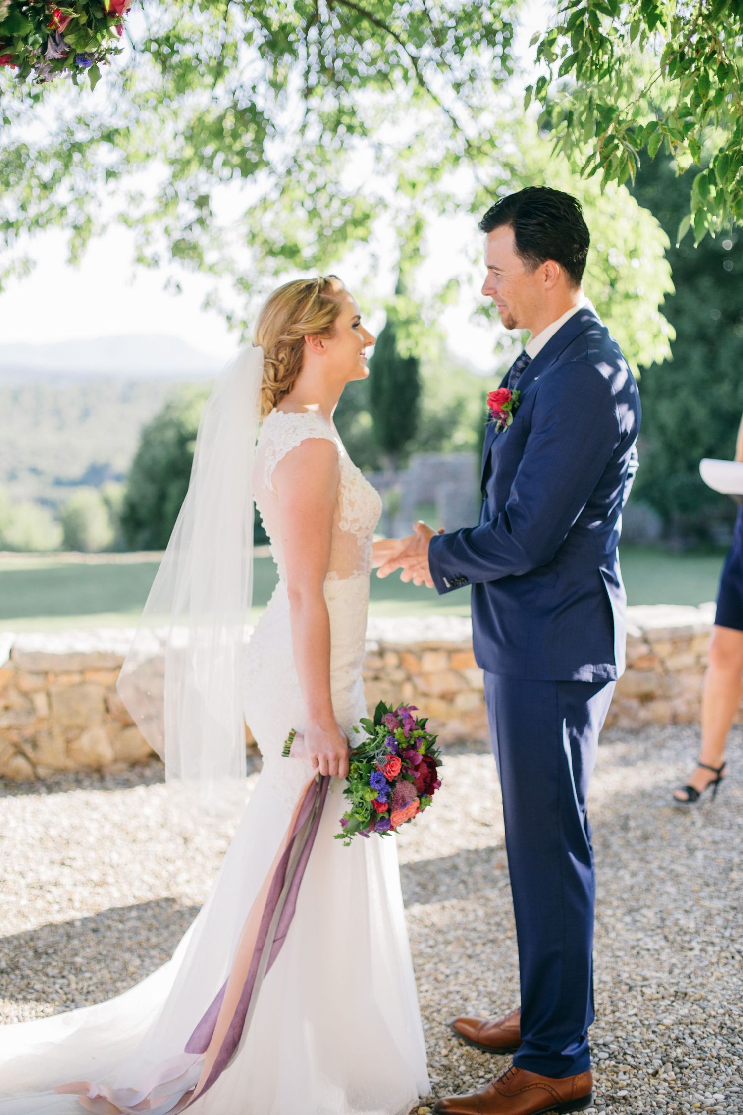 A bride in a white lace gown and groom in navy suit sharing a moment together outdoors.