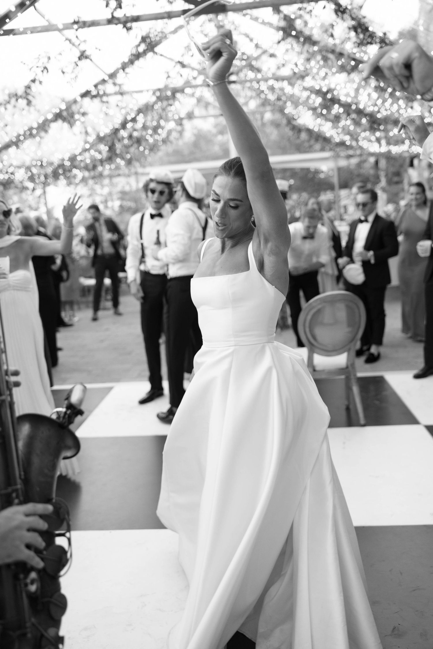 Bridesmaids in white dresses dancing with raised arms under a twinkling tent ceiling.