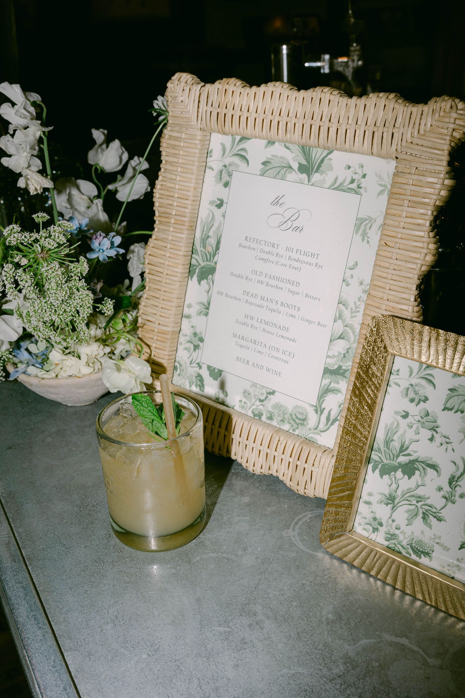 Wedding invitations with green botanical prints displayed in wicker frames alongside floral centerpieces.