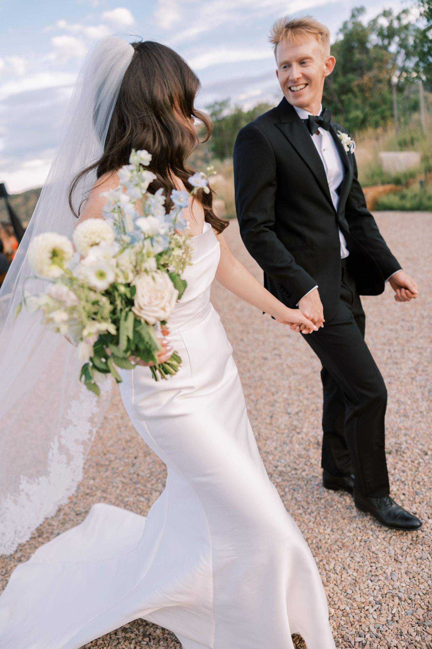 Elegant bride in white dress and groom in black tuxedo walk together on gravel path during outdoor wedding ceremony.