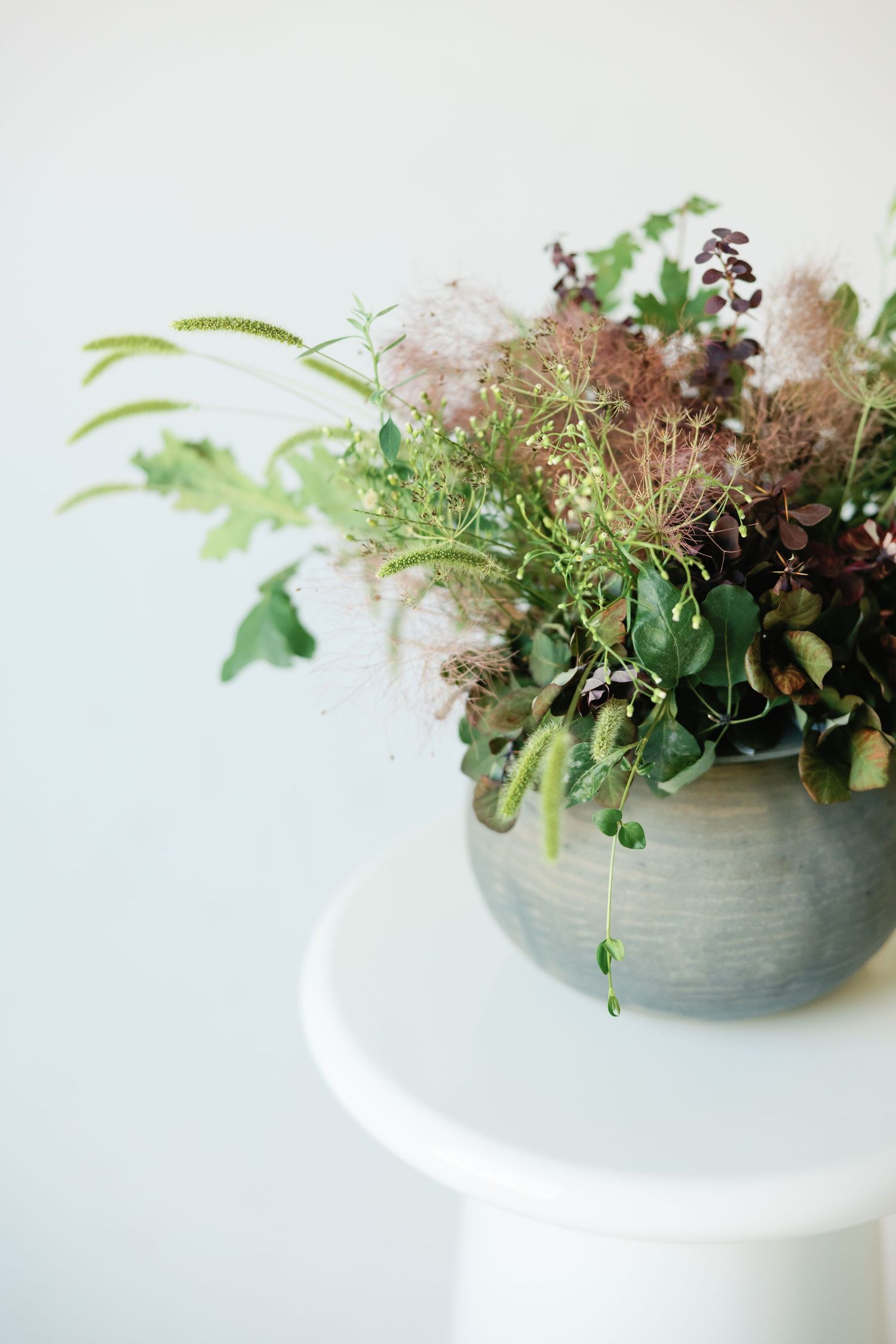 A ceramic bowl filled with wild greenery and dried flowers on a white pedestal against a light background.